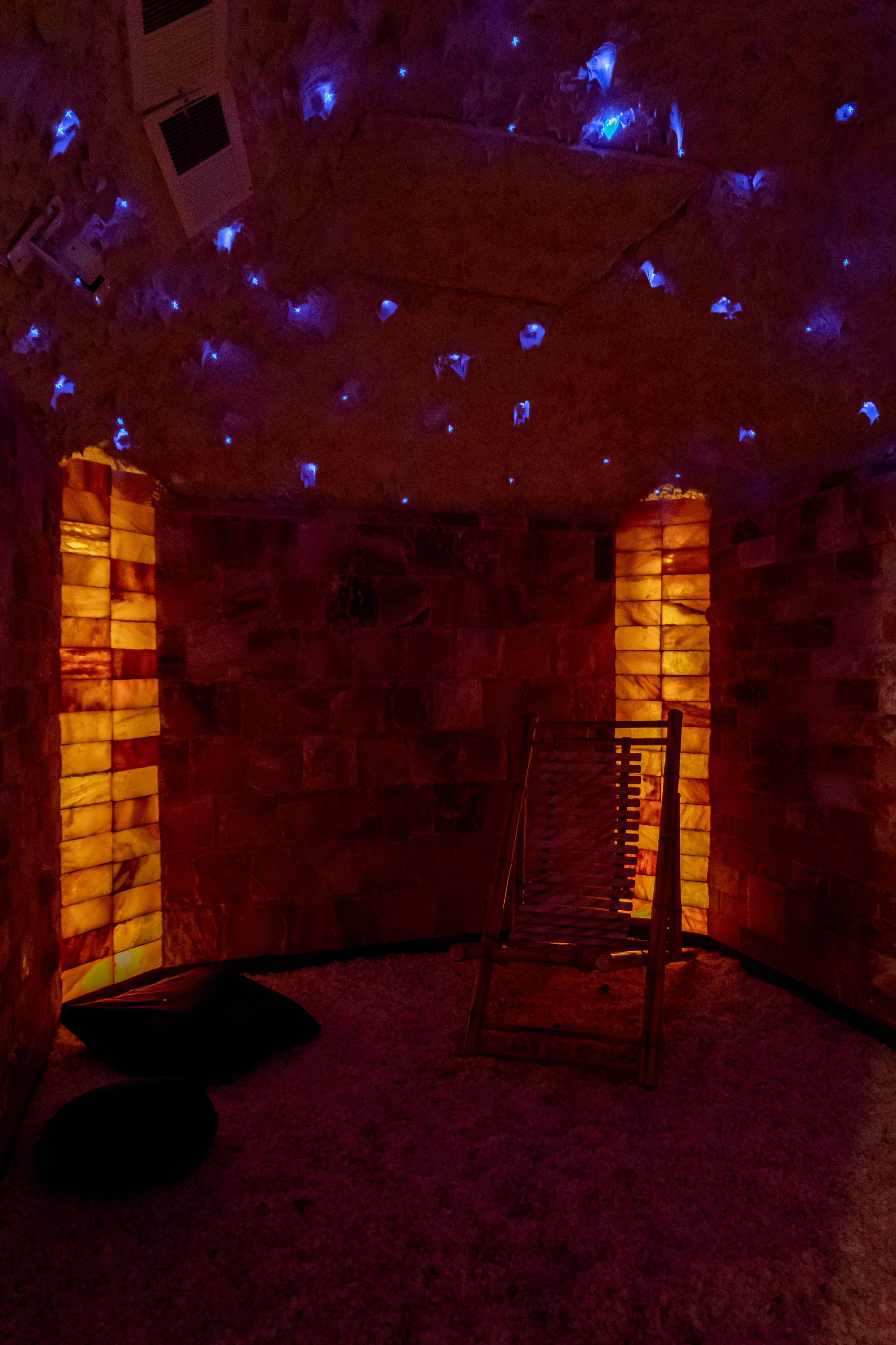 The image shows a dimly lit, circular salt therapy room with illuminated salt walls and a wooden reclining chair.