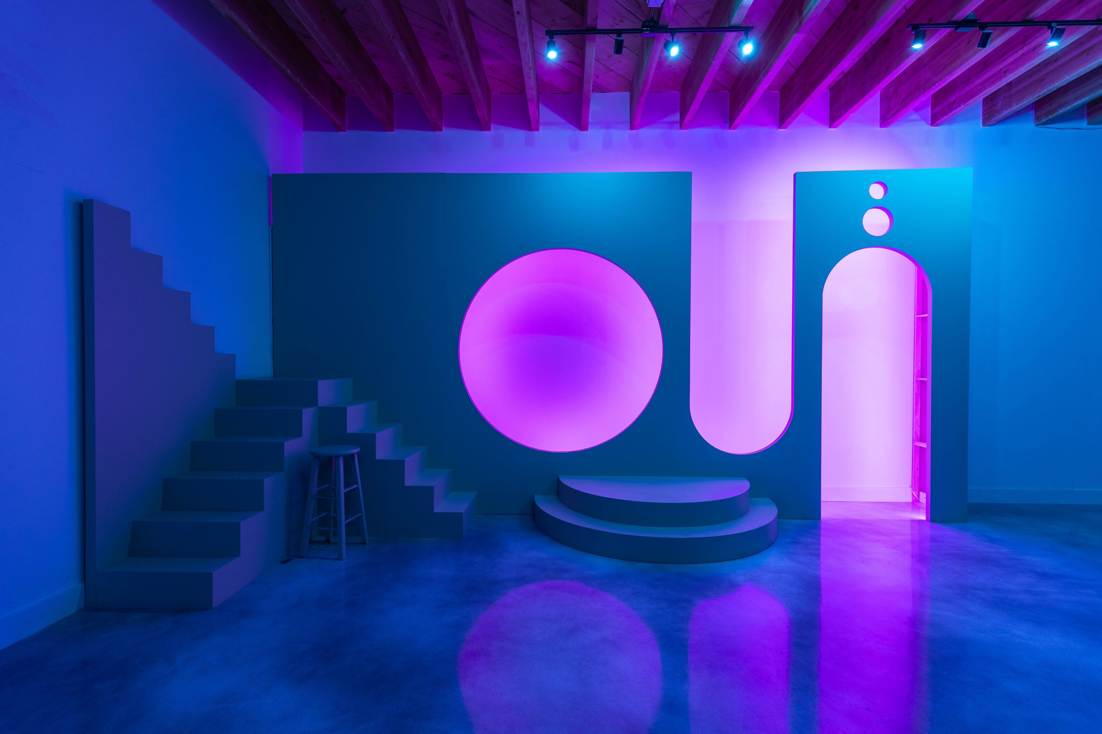 The image shows a modern interior with abstract shapes, featuring a set of stairs, circular and curved structures, and illuminated in shades of blue and pink.