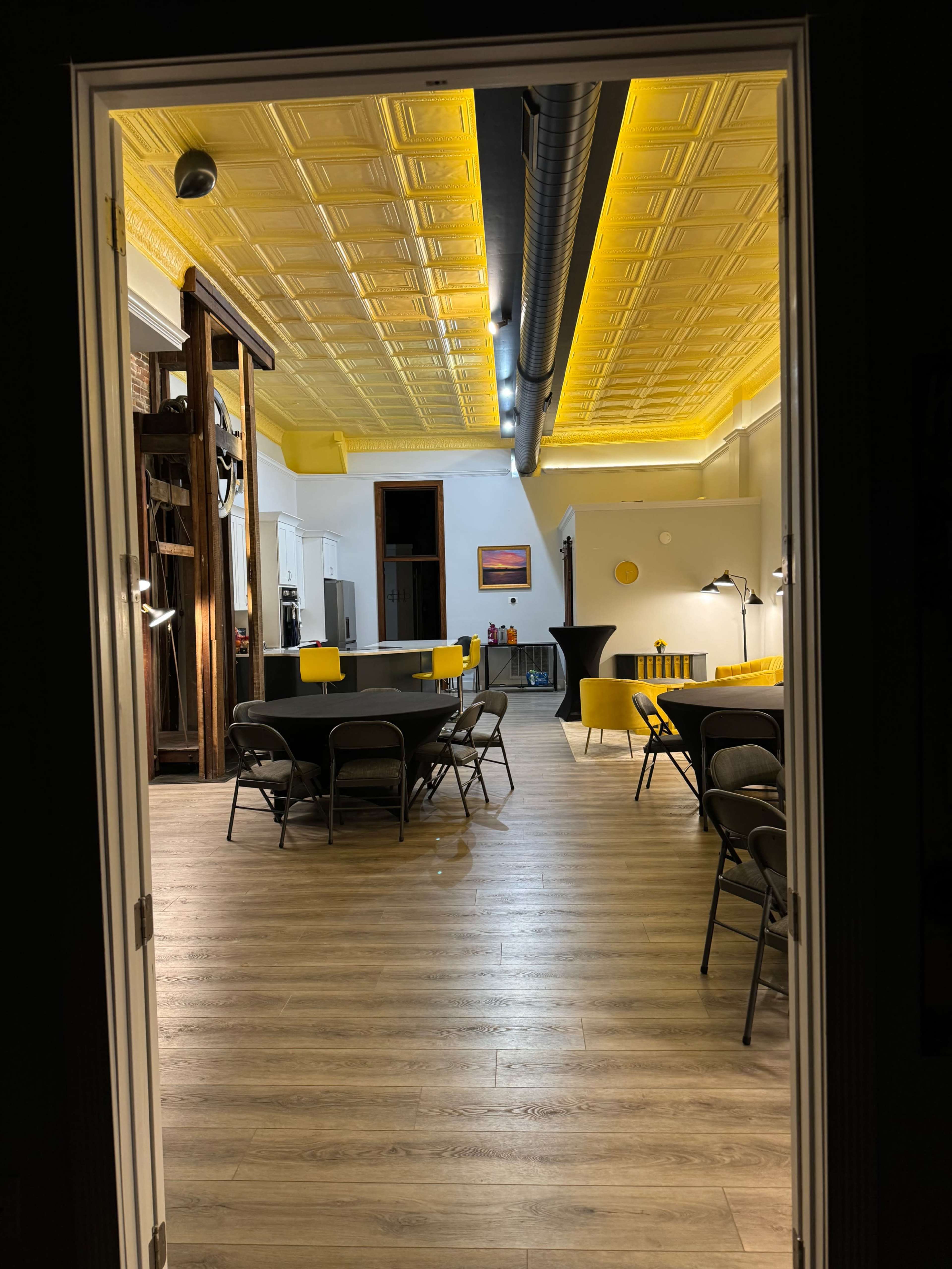 The image shows a spacious room with a yellow ceiling, a few tables and chairs, and kitchen appliances visible in the background.