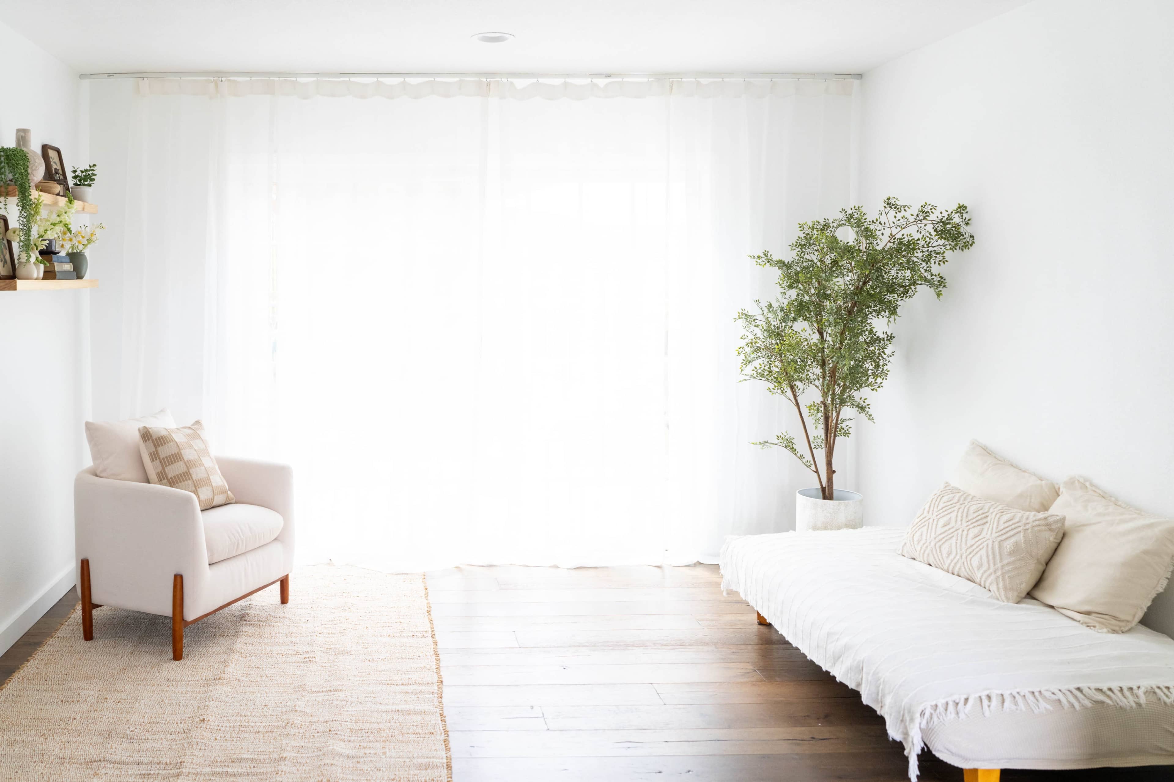 The image shows a bright, minimalistic room featuring a light-colored armchair, a sofa with decorative cushions, a small tree in a pot, and sheer curtains filtering sunlight.