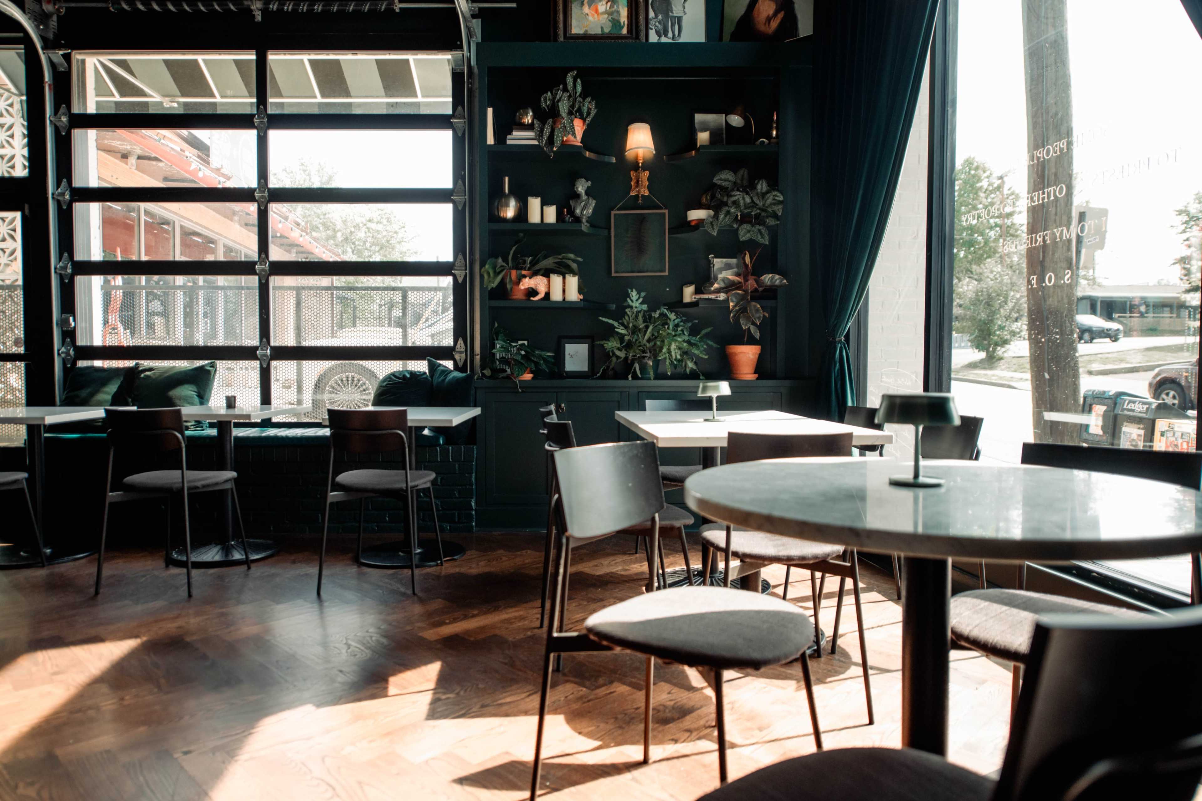 A modern café features dark green walls adorned with plants and decorative items, as well as simple black chairs and round tables arranged on a wooden floor.
