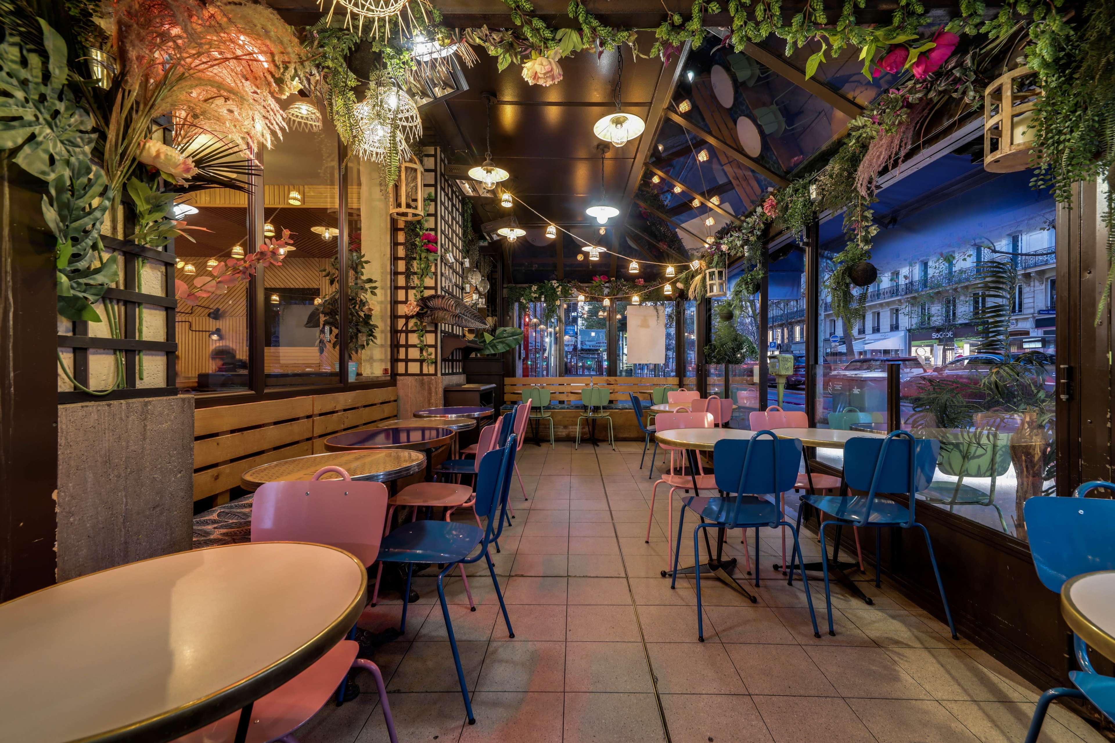The image shows a brightly decorated indoor café with colorful tables and chairs, surrounded by plants and string lights.