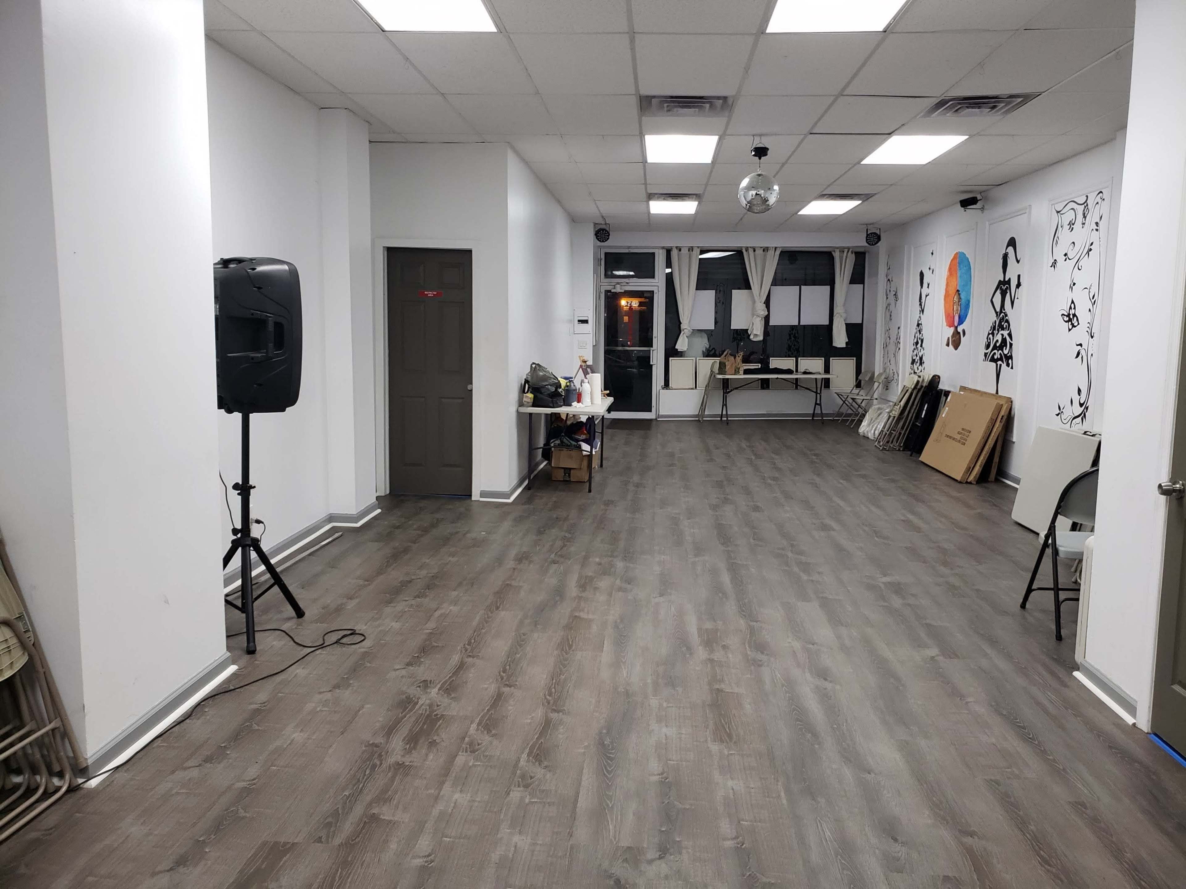 An empty interior space with wooden flooring, white walls, and a speaker on a stand near the entrance, along with various items stacked on tables.