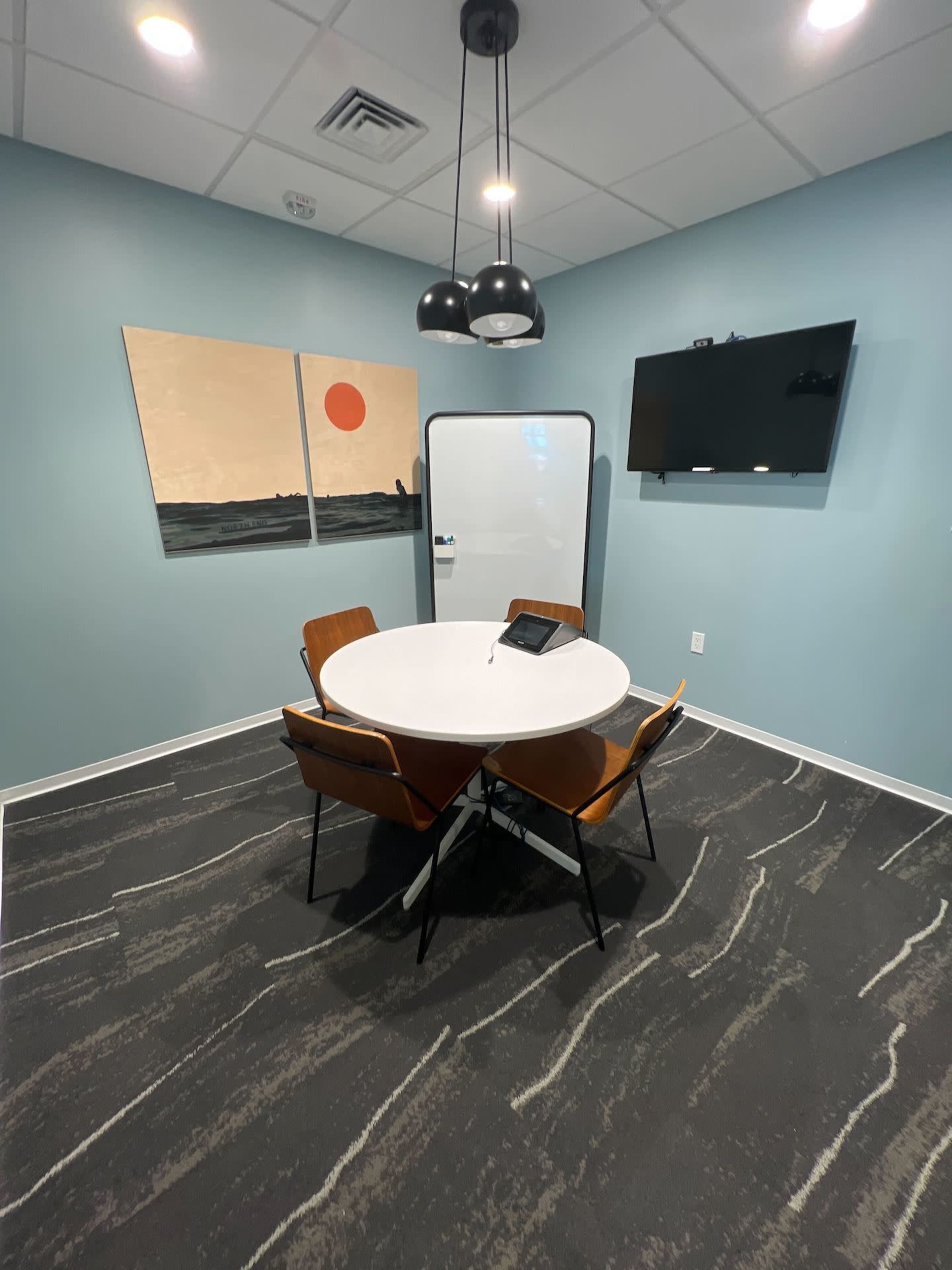 A small conference room features a round table, four chairs, a flat-screen TV, a whiteboard, and two abstract paintings on the wall.
