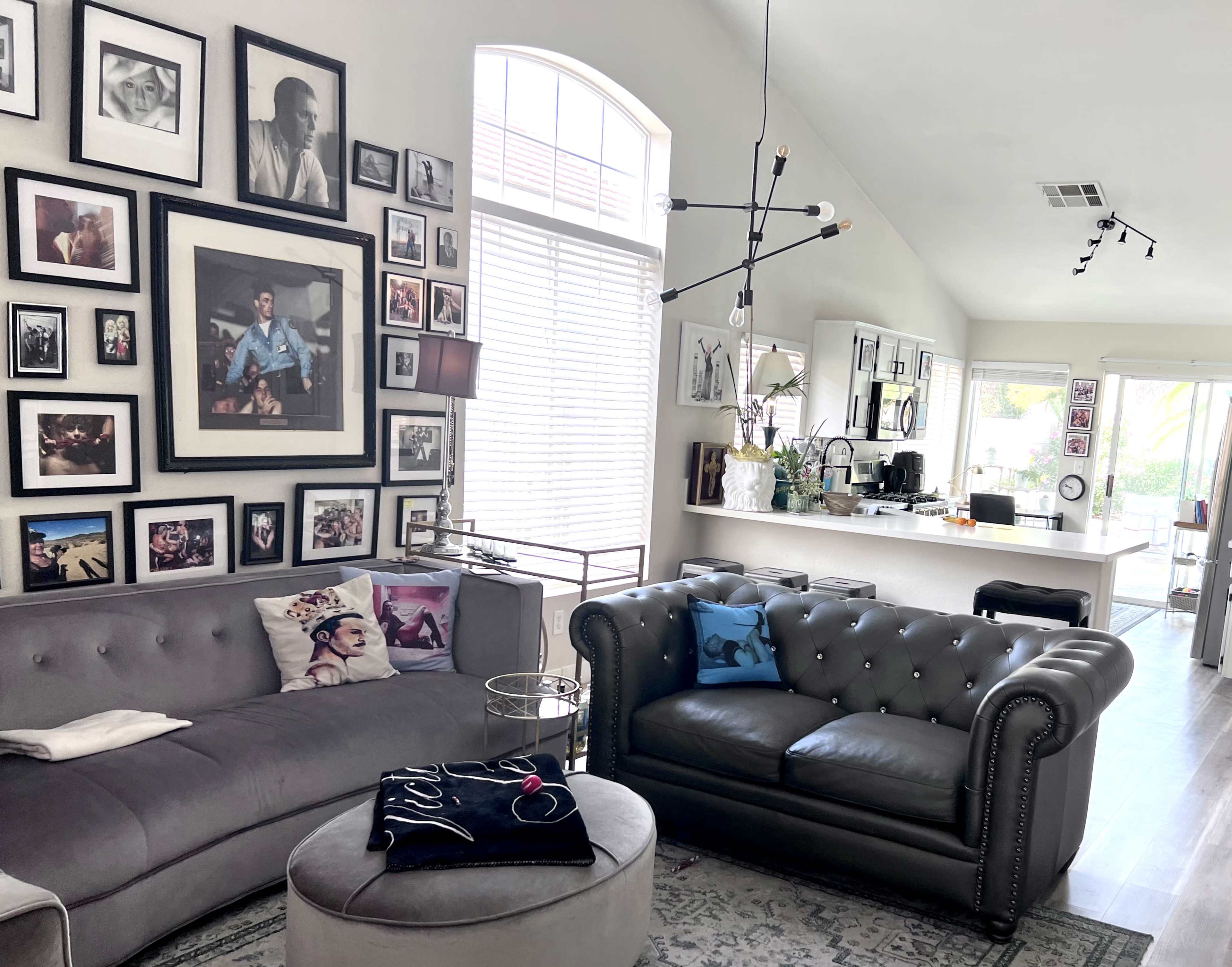 The living room features a gray sectional sofa and a black tufted armchair, surrounded by a gallery wall of framed photos and artwork, with a modern light fixture overhead.