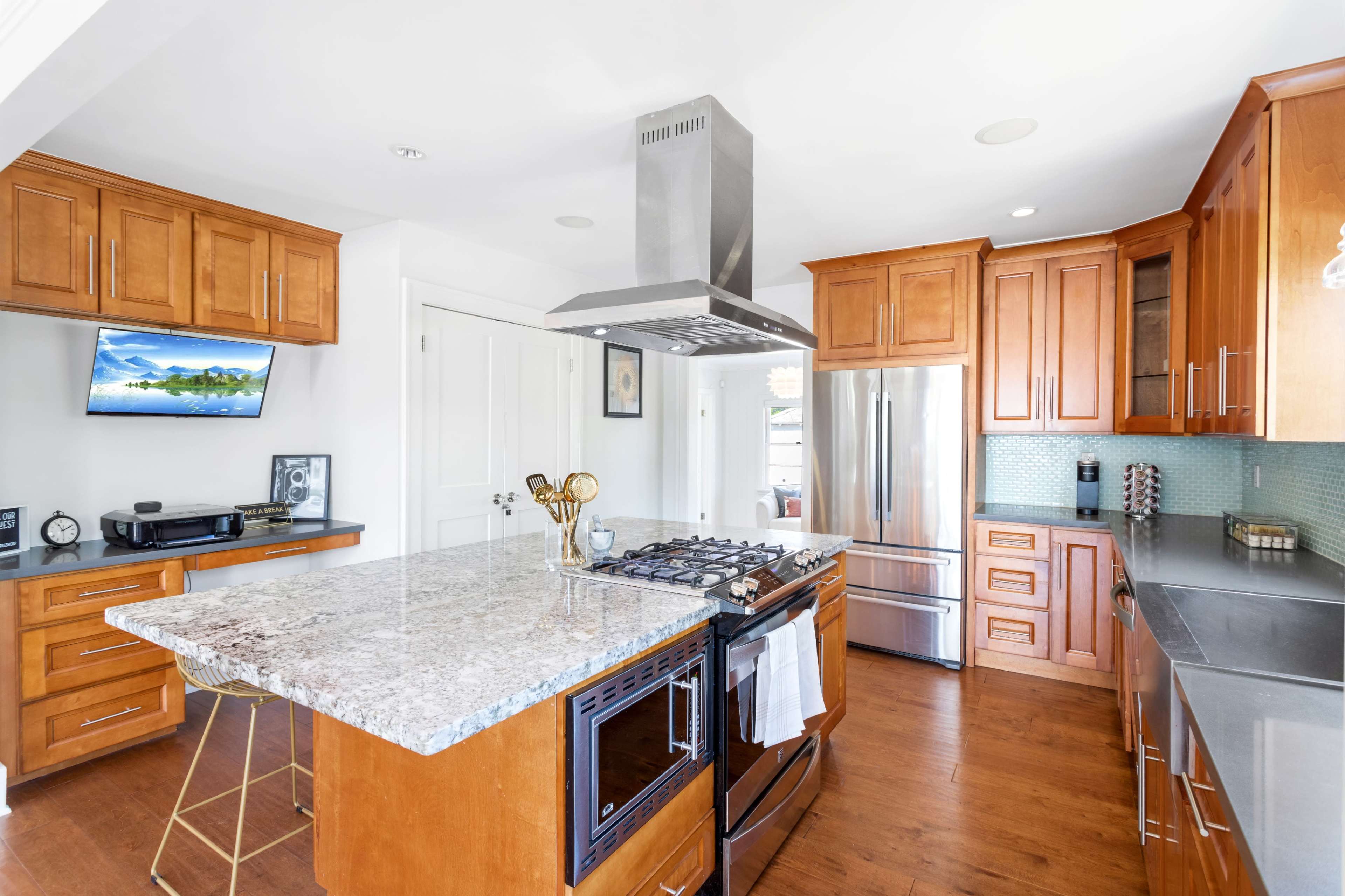 A modern kitchen with wooden cabinets, a granite island, stainless steel appliances, and a television mounted on the wall.