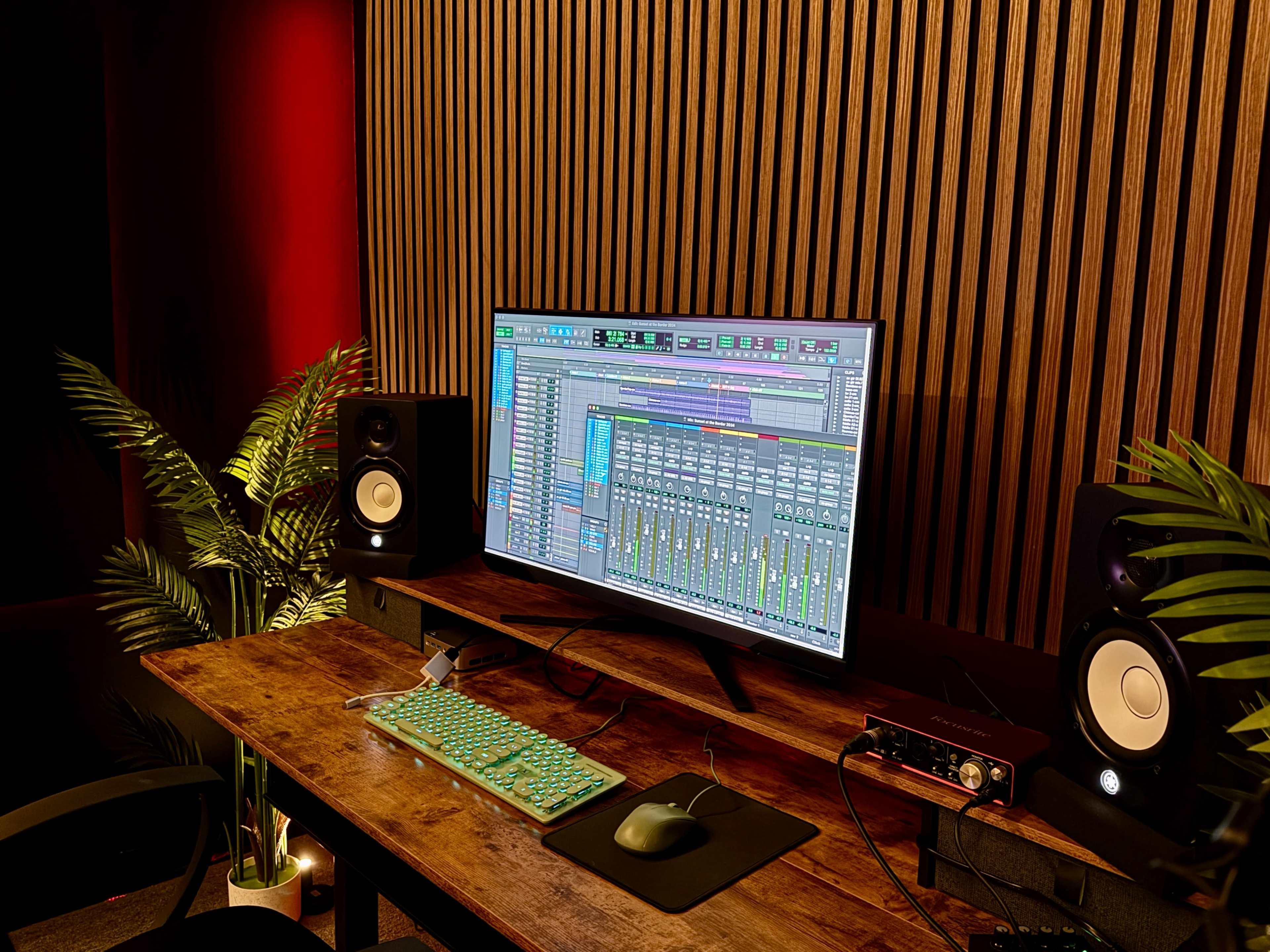 A music production workspace features a computer monitor displaying audio mixing software, flanked by studio monitors, with a wooden desk and decorative plants.