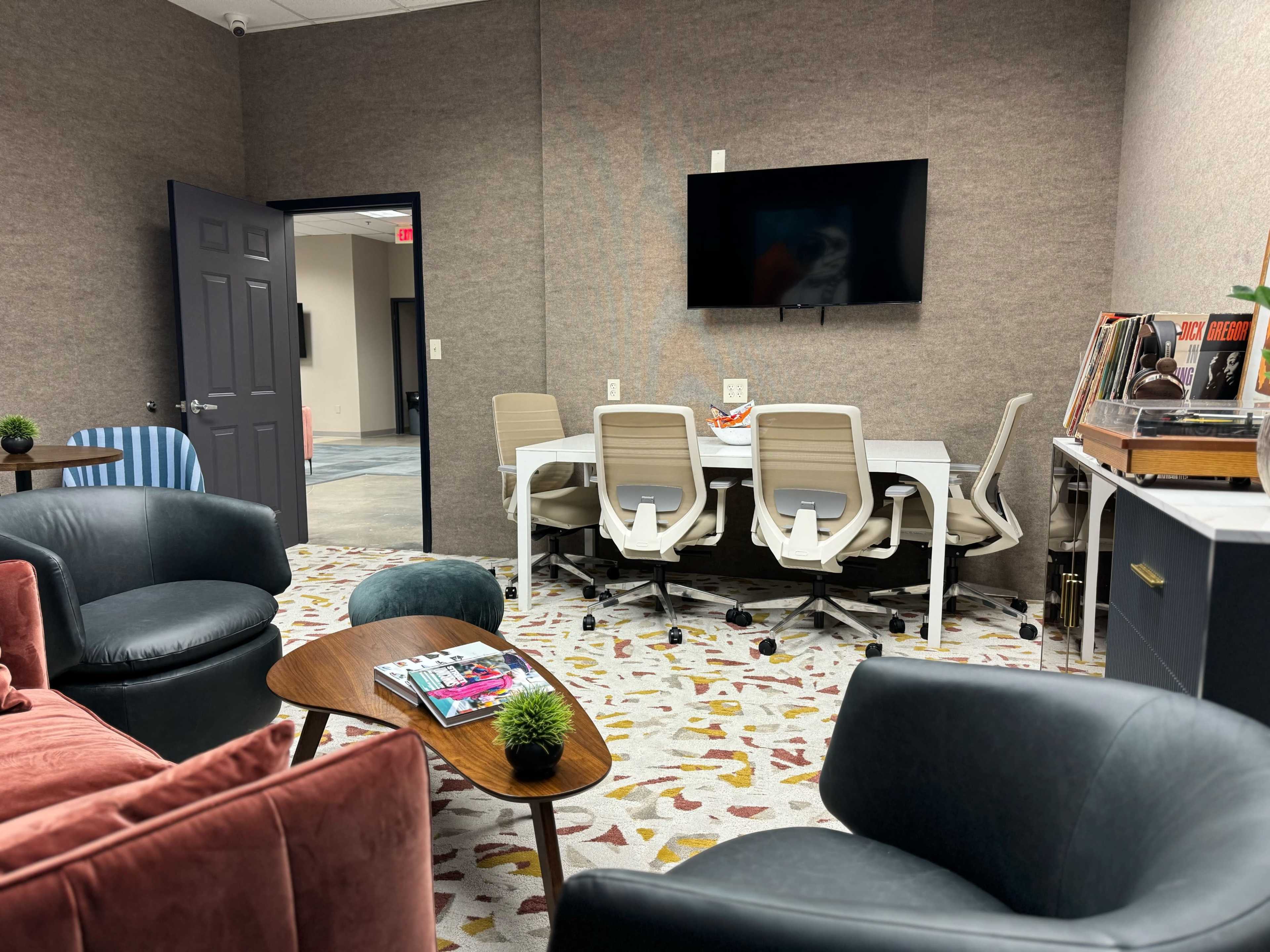A modern meeting room featuring a rectangular table surrounded by ergonomic chairs, a television mounted on the wall, and a lounge area with a sofa and armchairs.