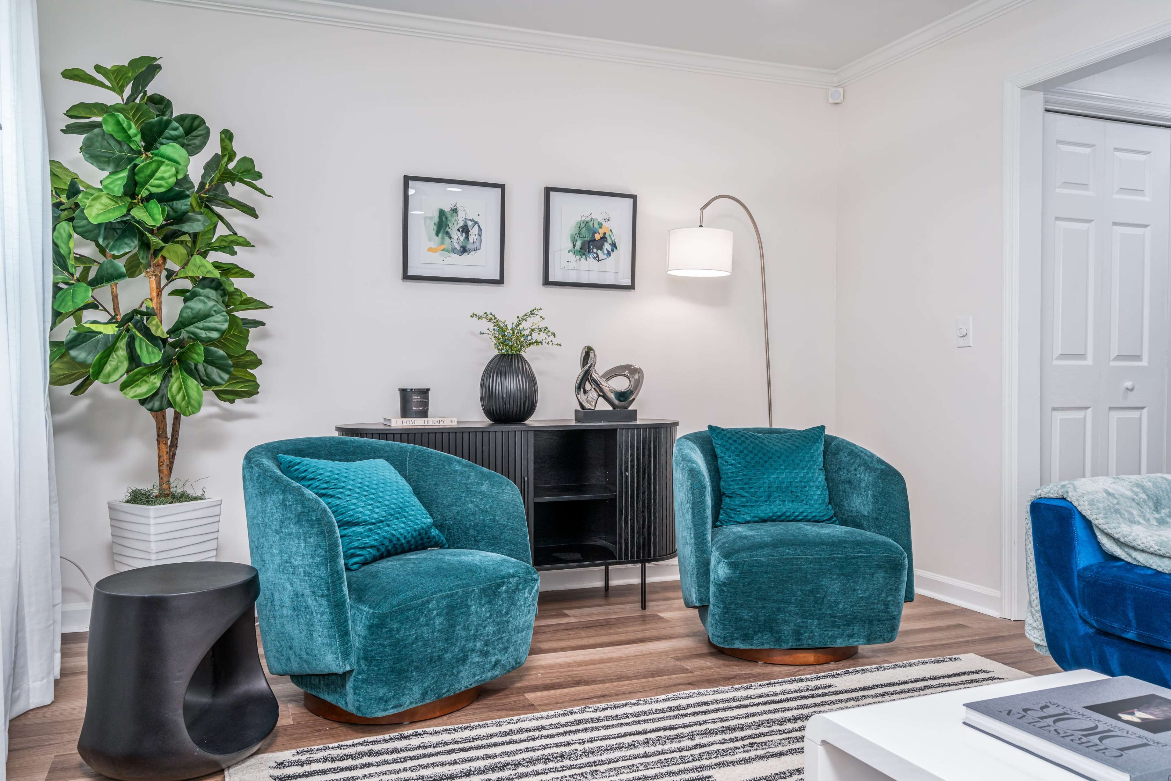 A modern living room with two teal chairs, a black side table, a cabinet, framed artwork, and a large indoor plant.