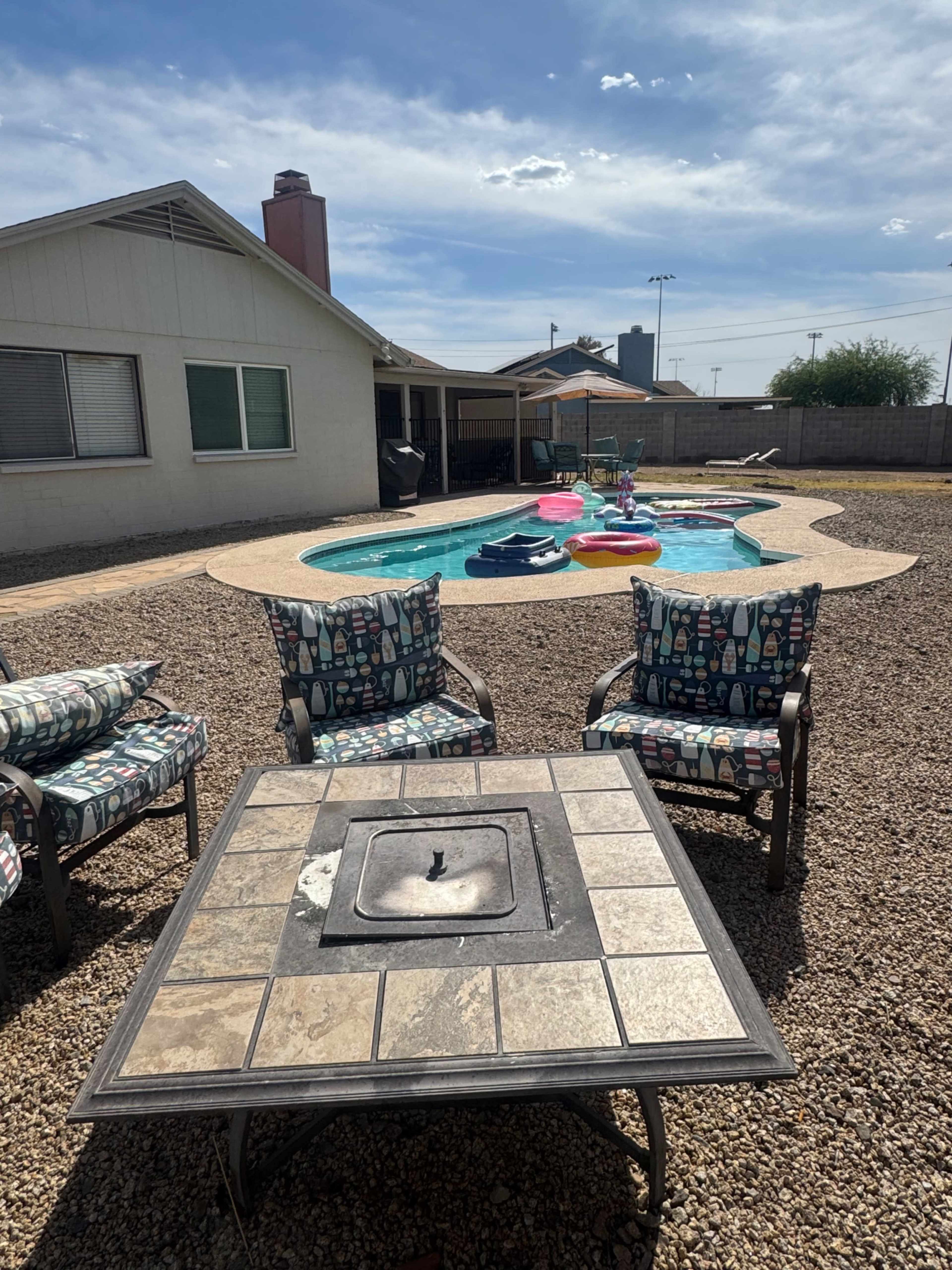 The image shows a backyard with a swimming pool, colorful floaties, and patio chairs arranged around a stone table.