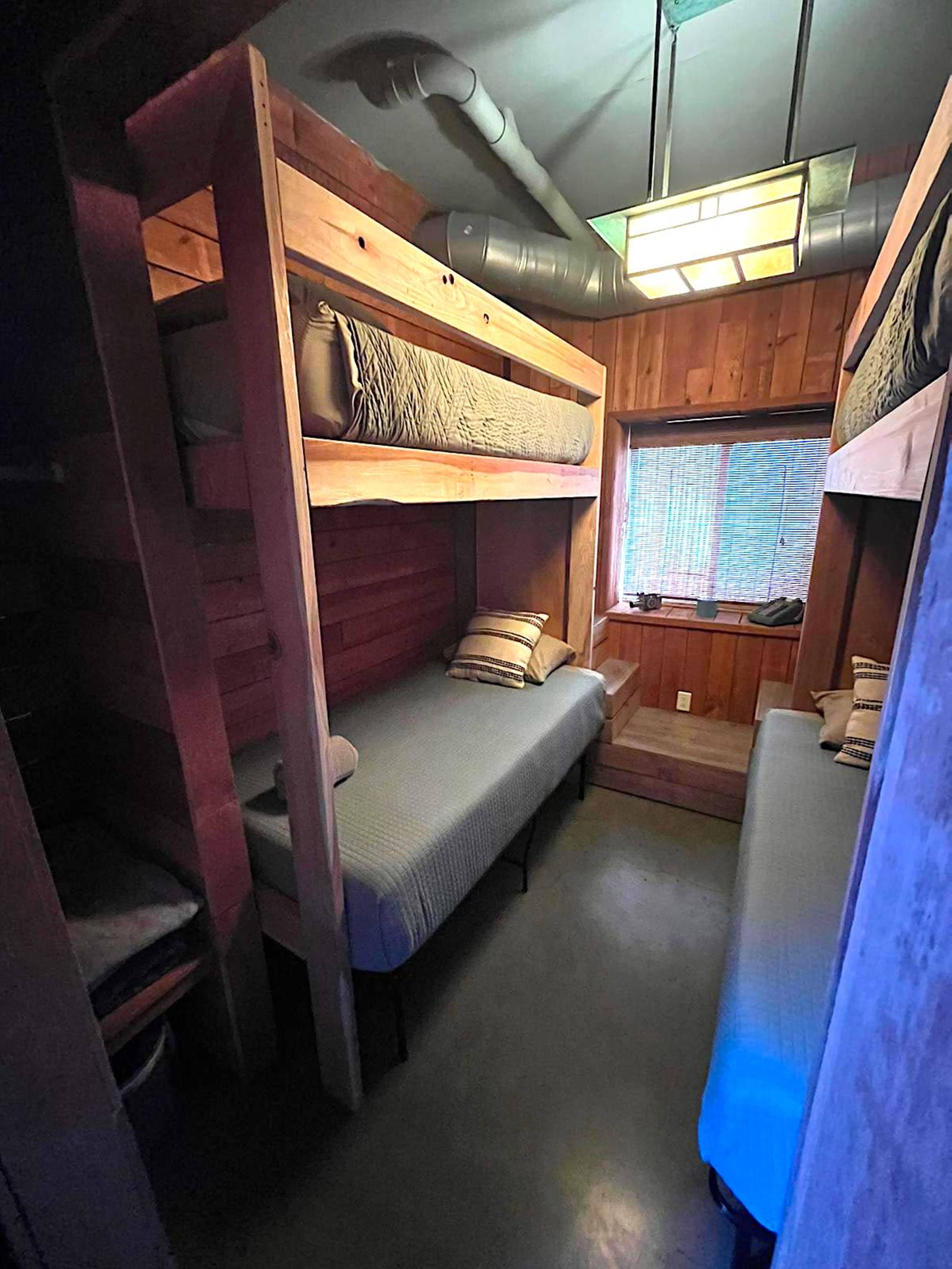 A small cabin room featuring two sets of wooden bunk beds and a window with a green light fixture overhead.