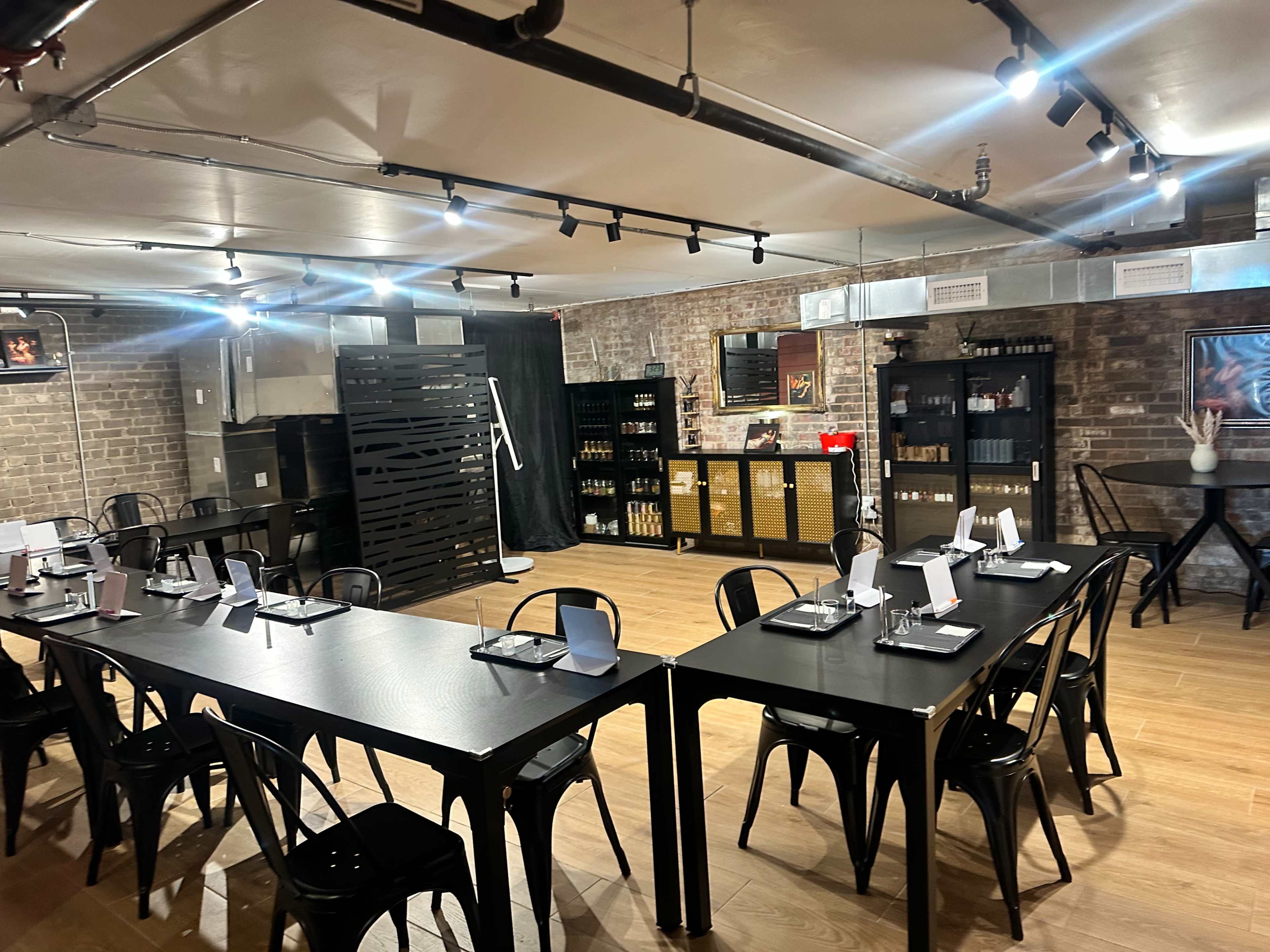 The image shows a modern dining area with several black tables and chairs arranged in an industrial-style space featuring exposed brick walls and shelves stocked with various items.