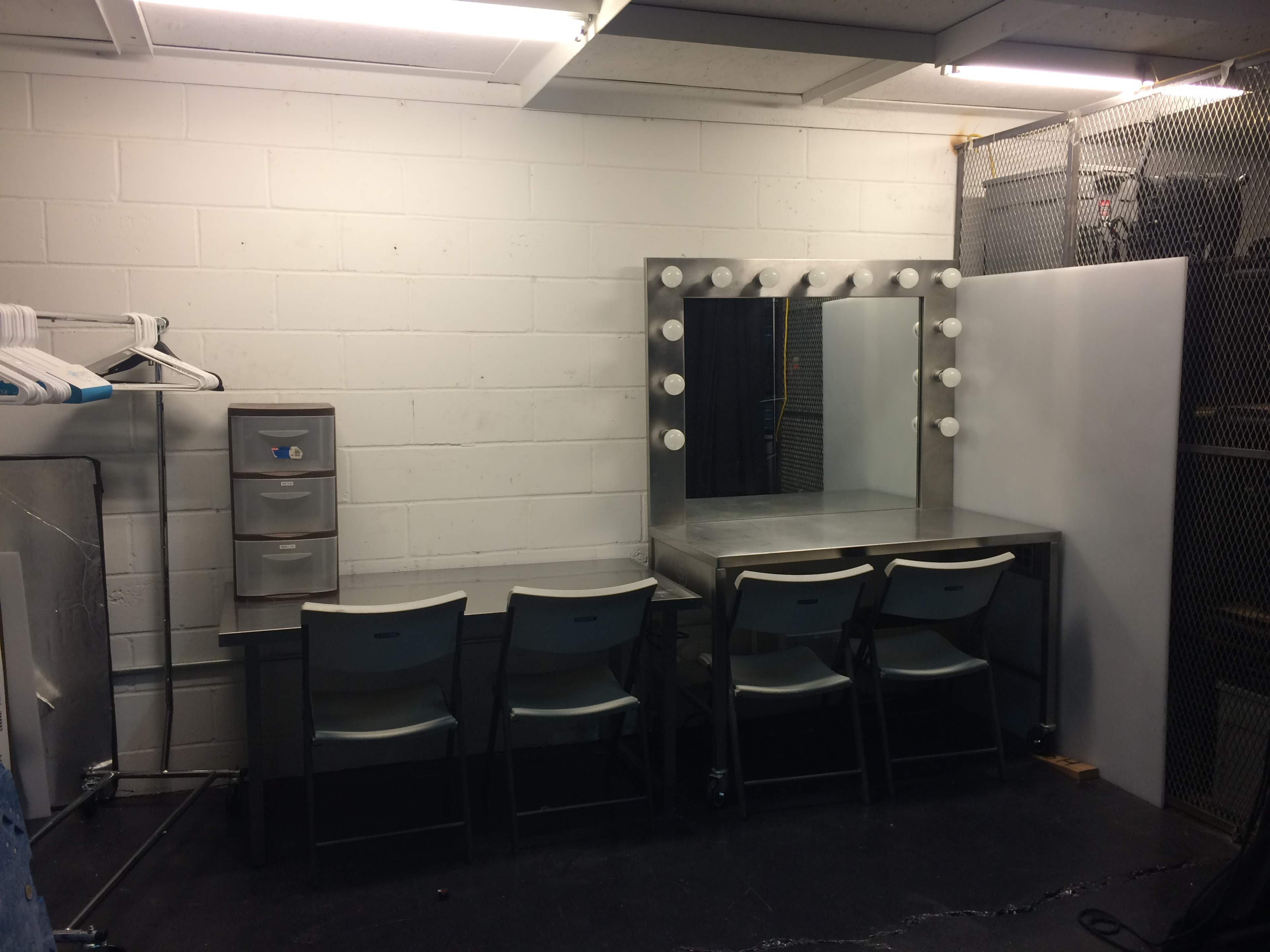 A simple backstage dressing room with a mirror surrounded by light bulbs, a metal table, and four plastic chairs.