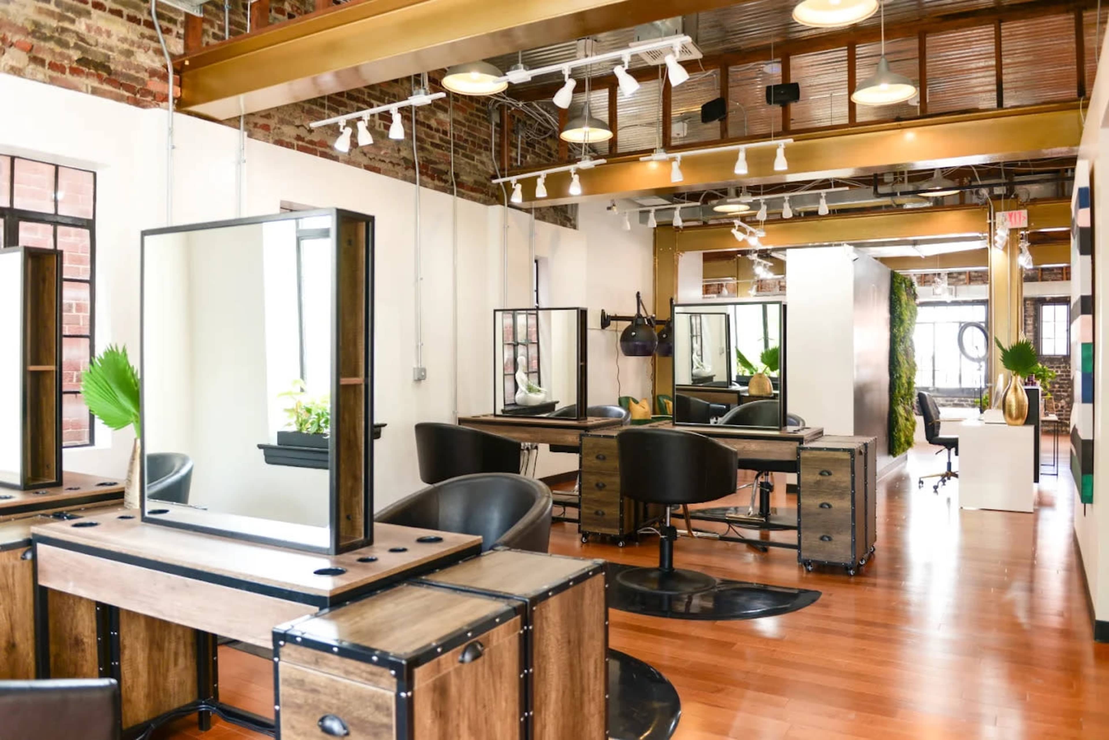 The image shows a modern hair salon featuring wooden styling stations with mirrors, black salon chairs, and large windows allowing natural light in.