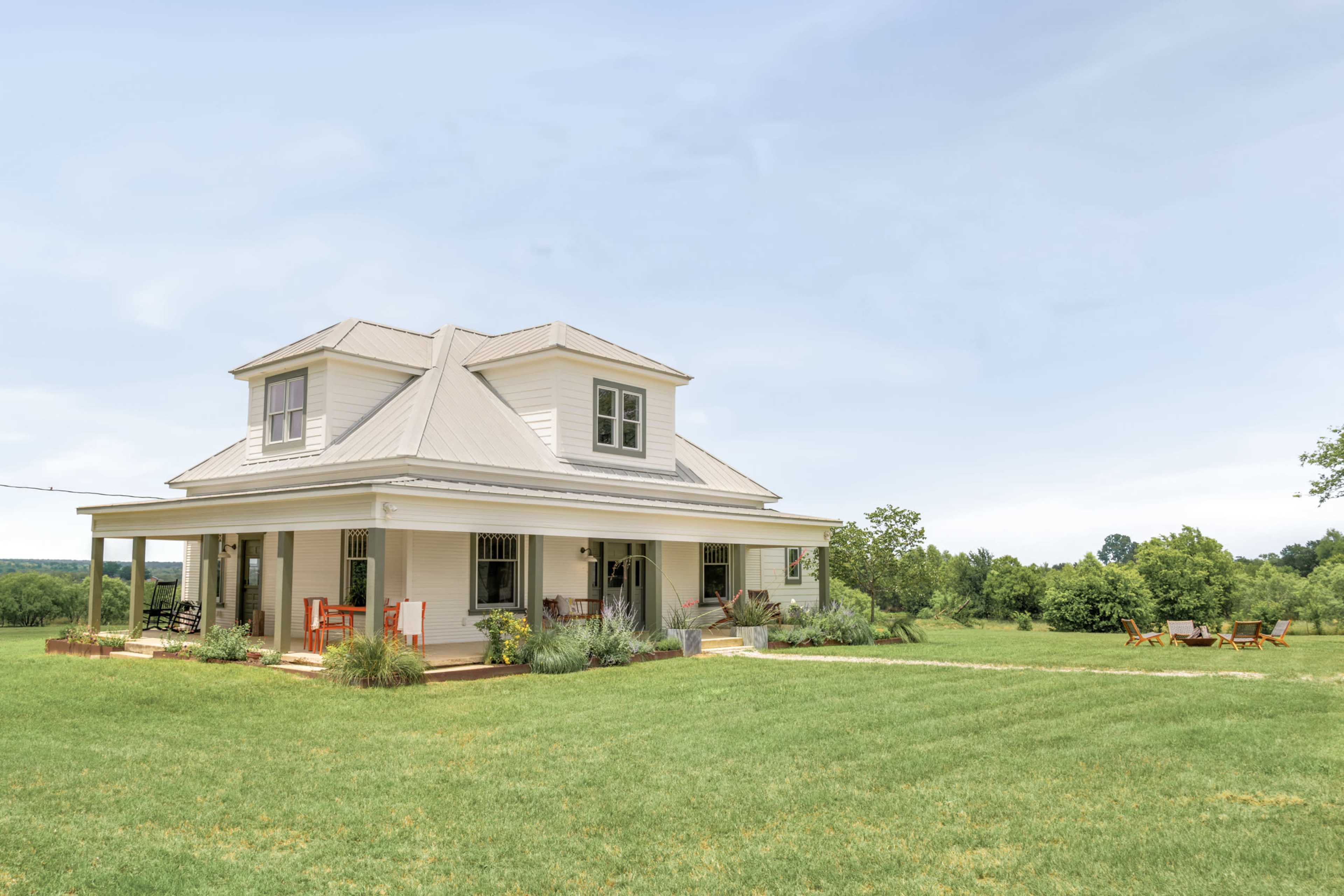 A white two-story house with a metal roof is surrounded by a green lawn and small trees in an open field.