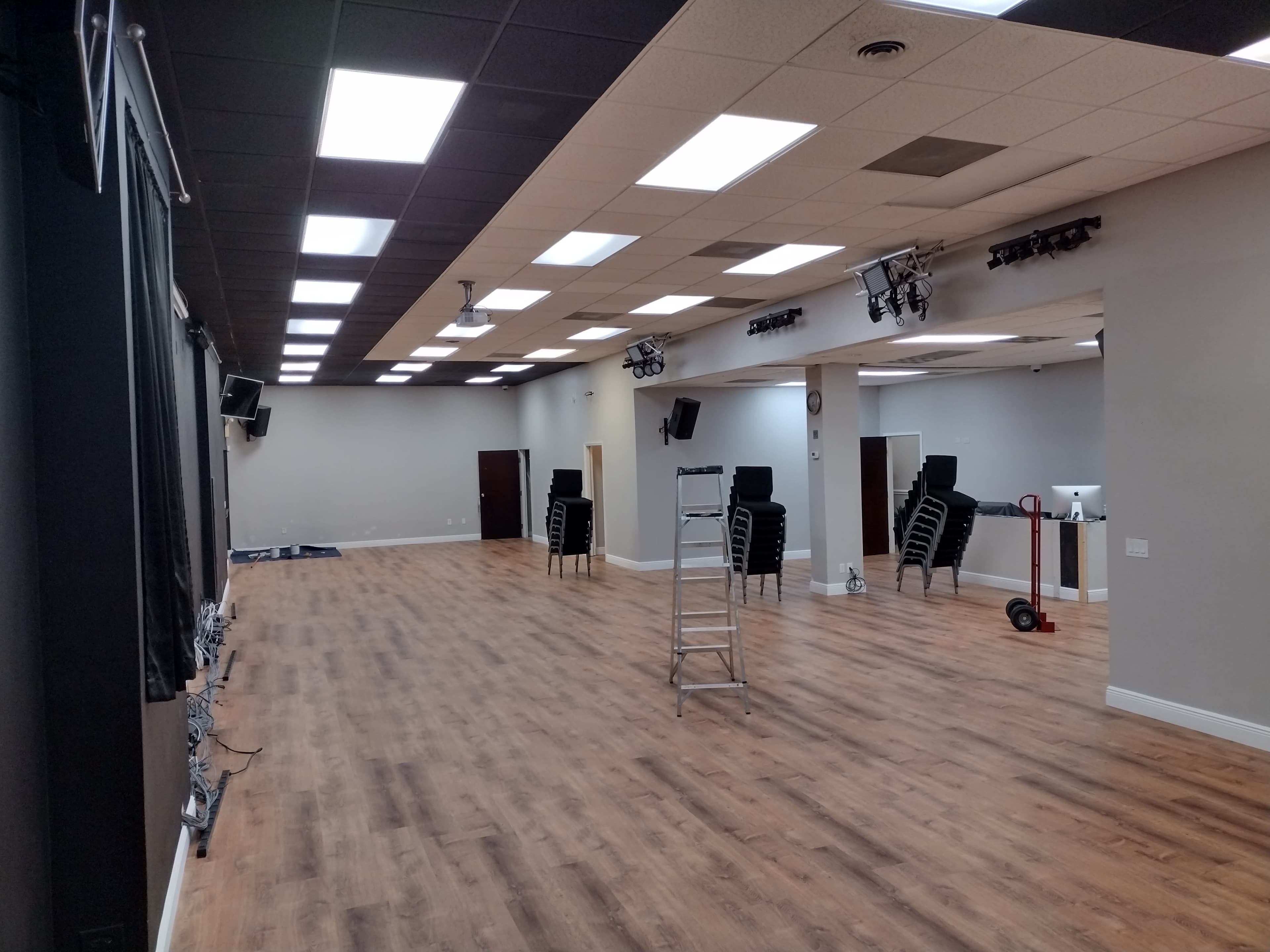 An empty, well-lit indoor space with wooden flooring, several stacked chairs, and a ladder positioned in the center.