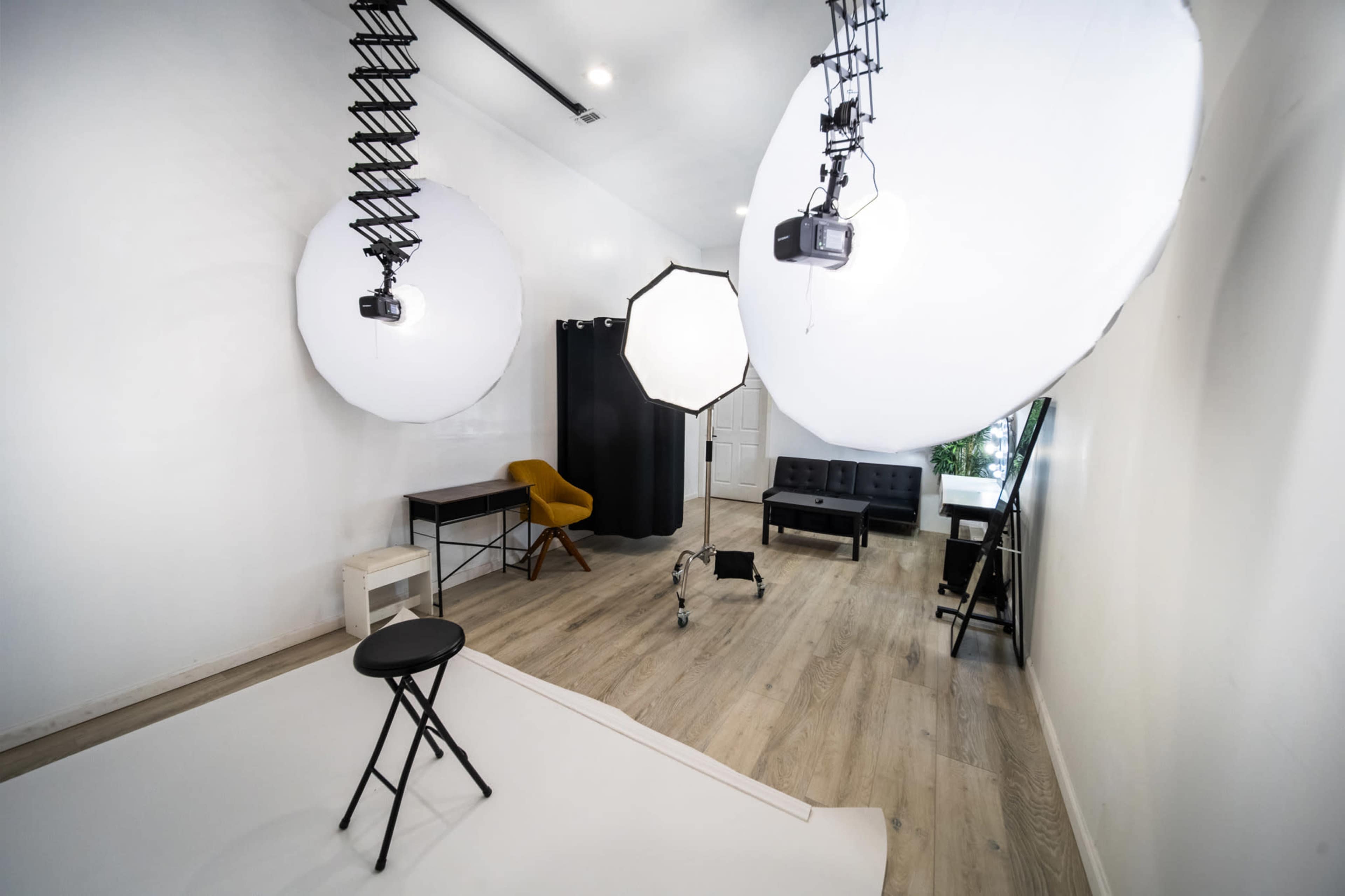 The image shows a photography studio featuring softbox lighting setups, a stool, two dark sofas, and a backdrop curtain arranged in a well-lit space.