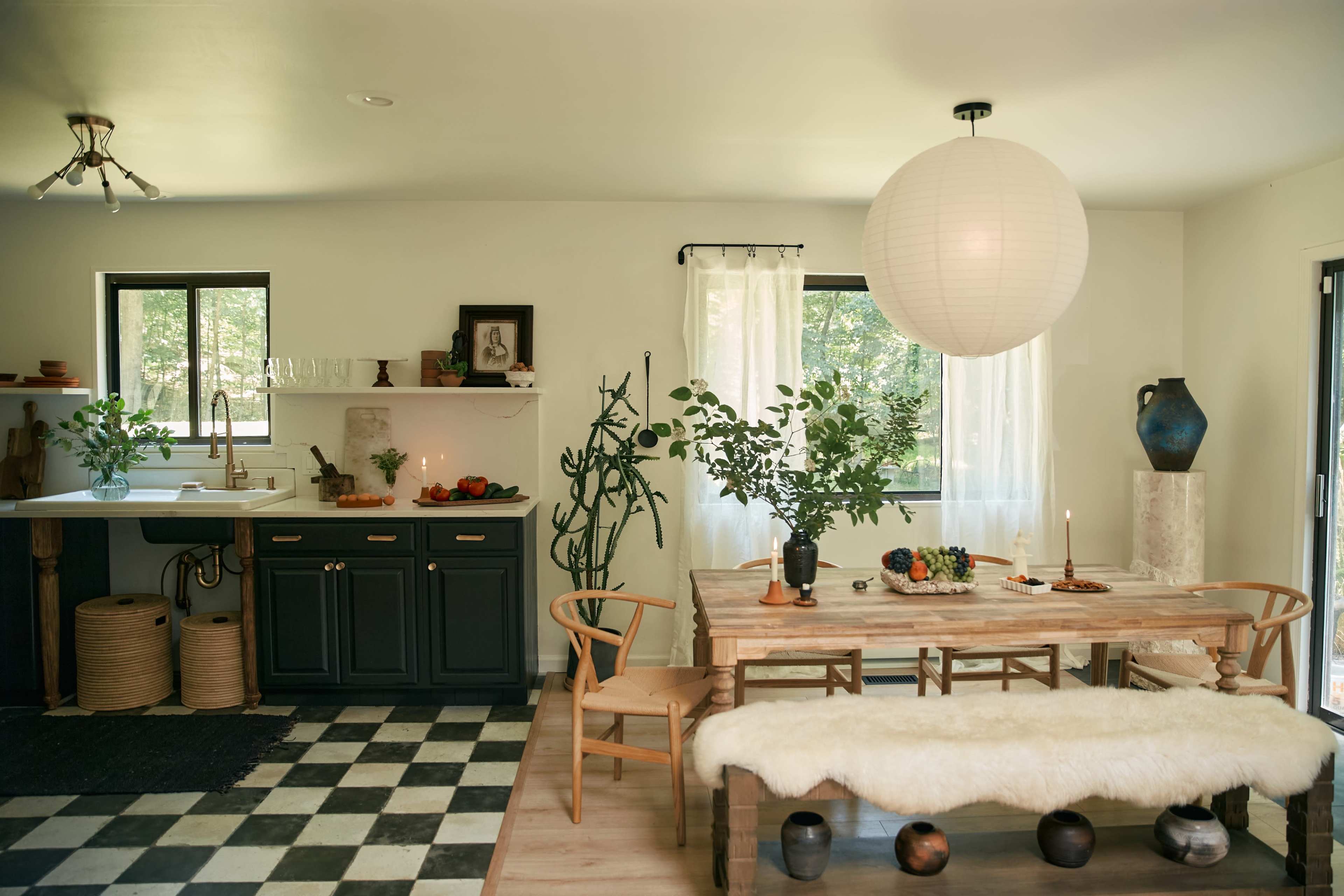 A modern dining area features a wooden table with a centerpiece, surrounded by chairs, and a kitchen in the background with dark cabinetry and a window revealing greenery outside.