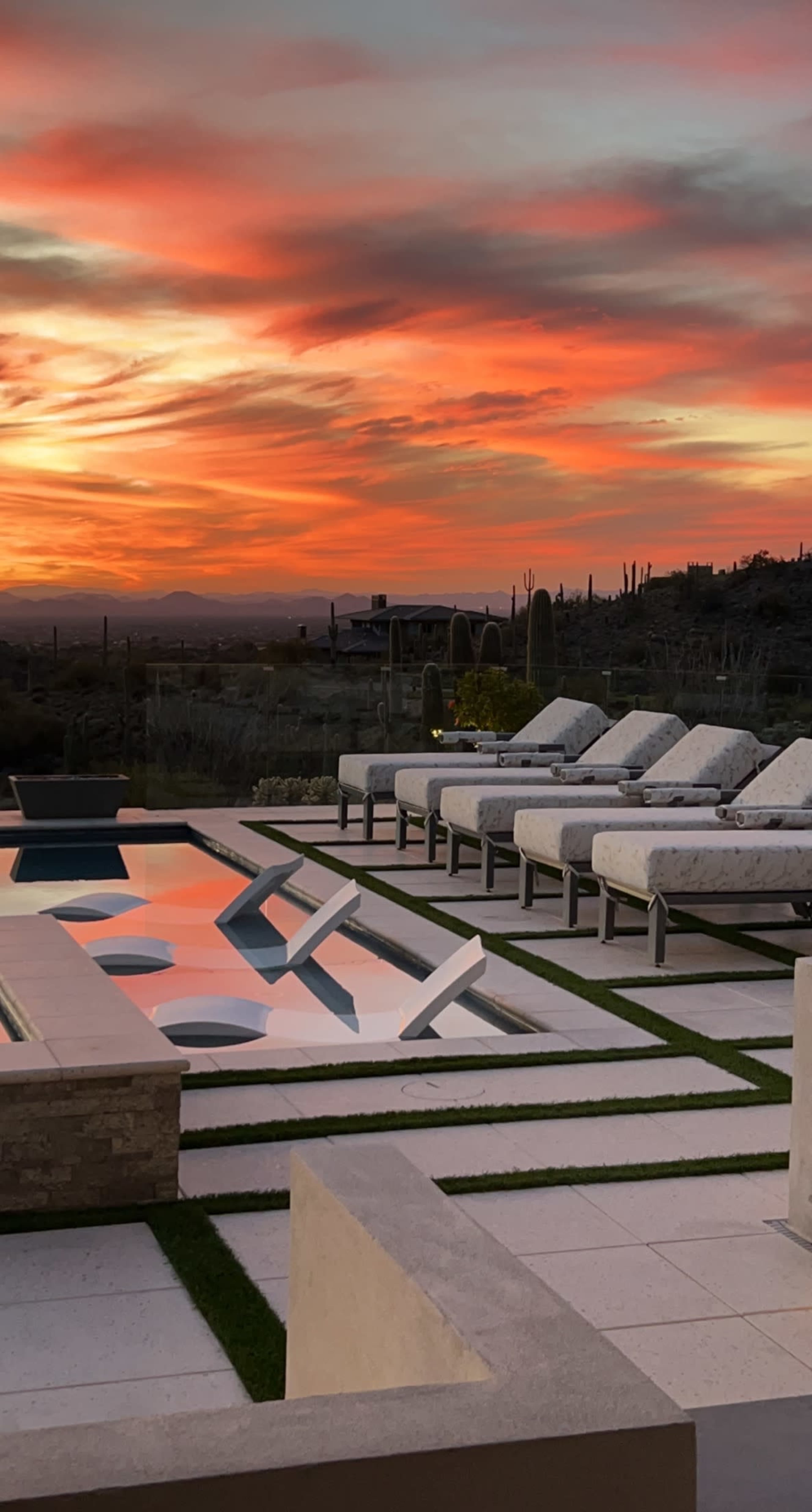 The scene shows a modern outdoor patio with lounge chairs and a pool reflecting a vibrant sunset sky.