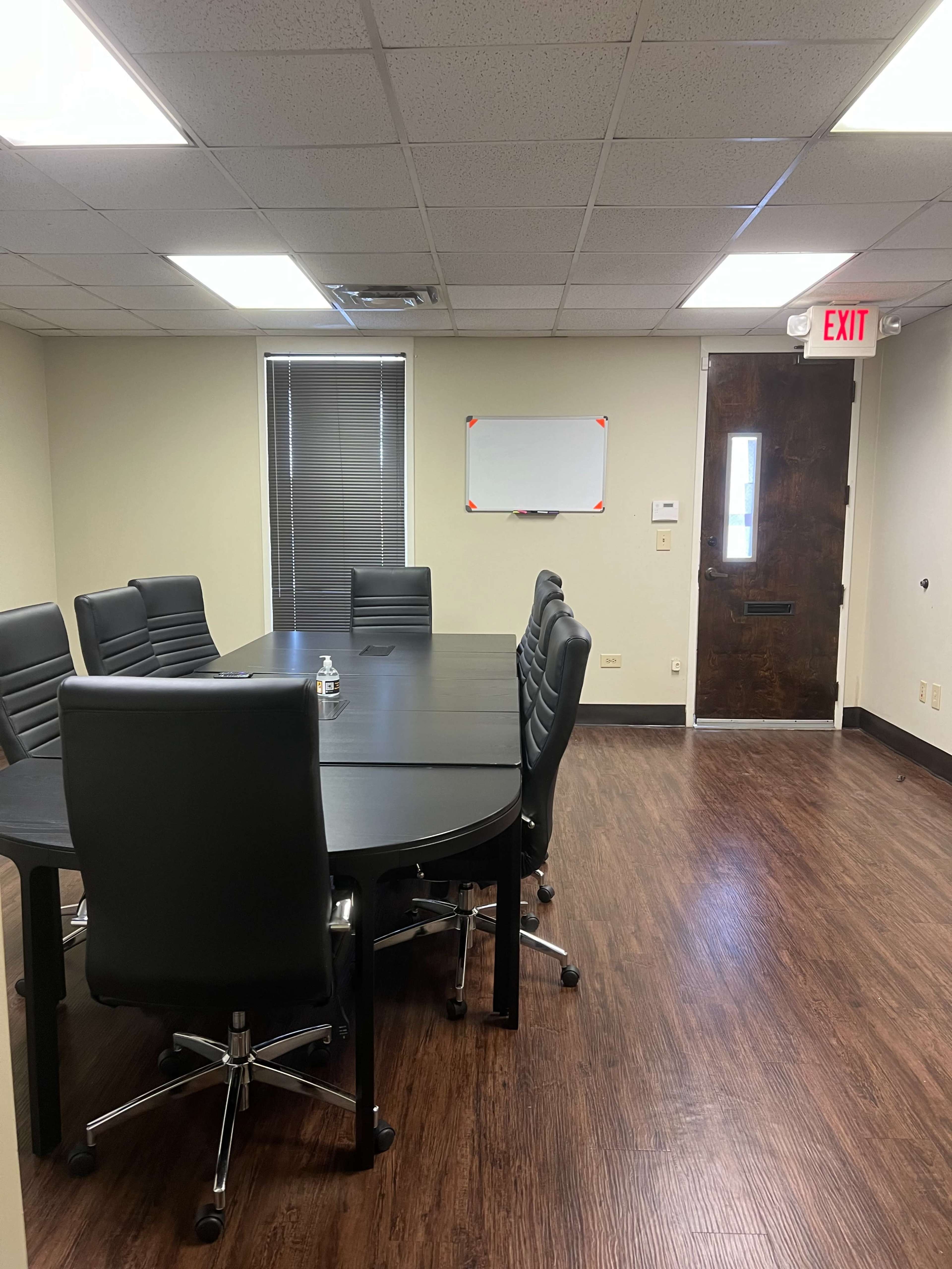 A conference room features a large, oval table surrounded by black swivel chairs, with a whiteboard and exit door visible in the background.