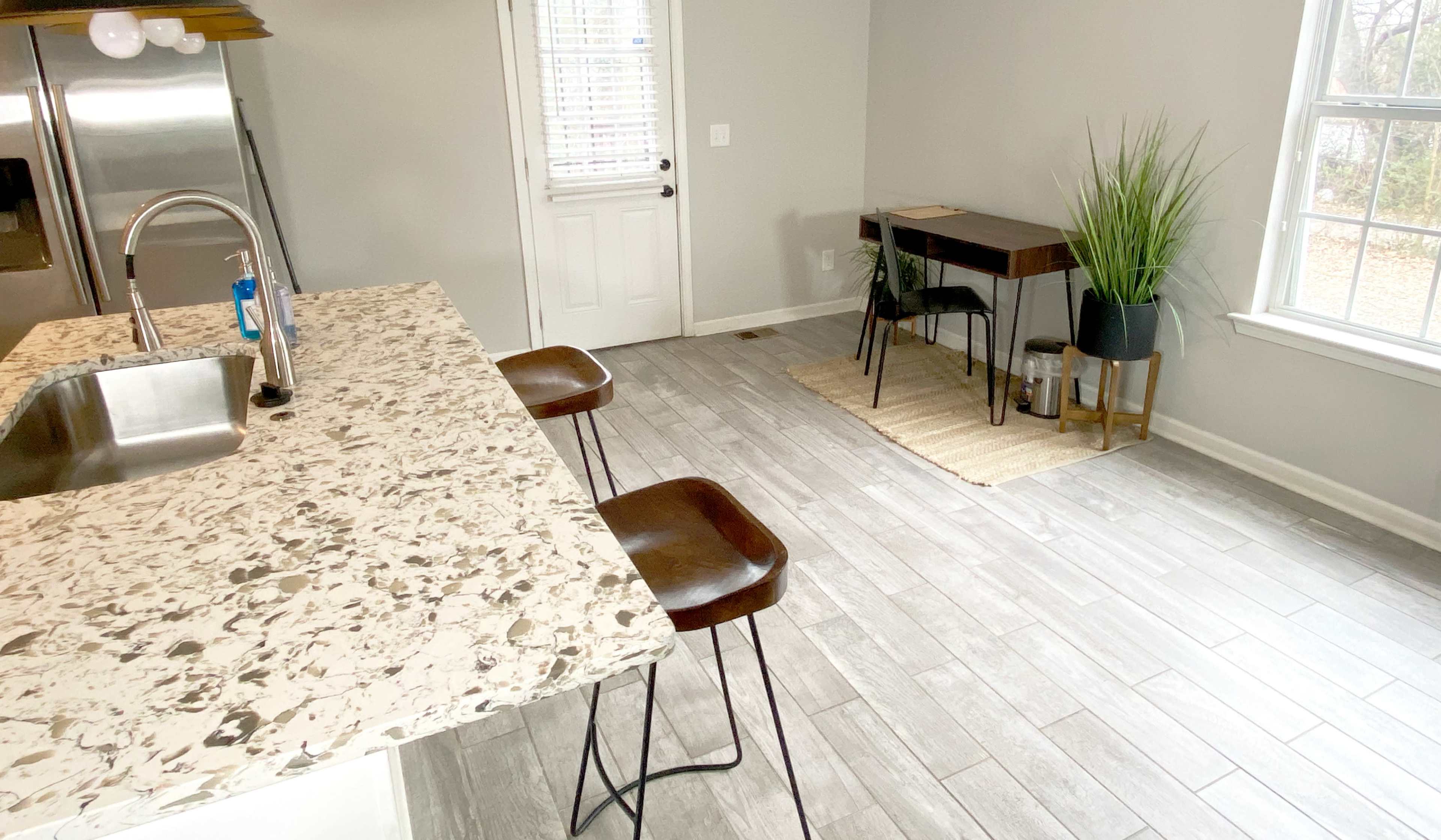 The image shows a kitchen area with a bar counter and two stools, alongside a small workspace and a window with a potted plant.