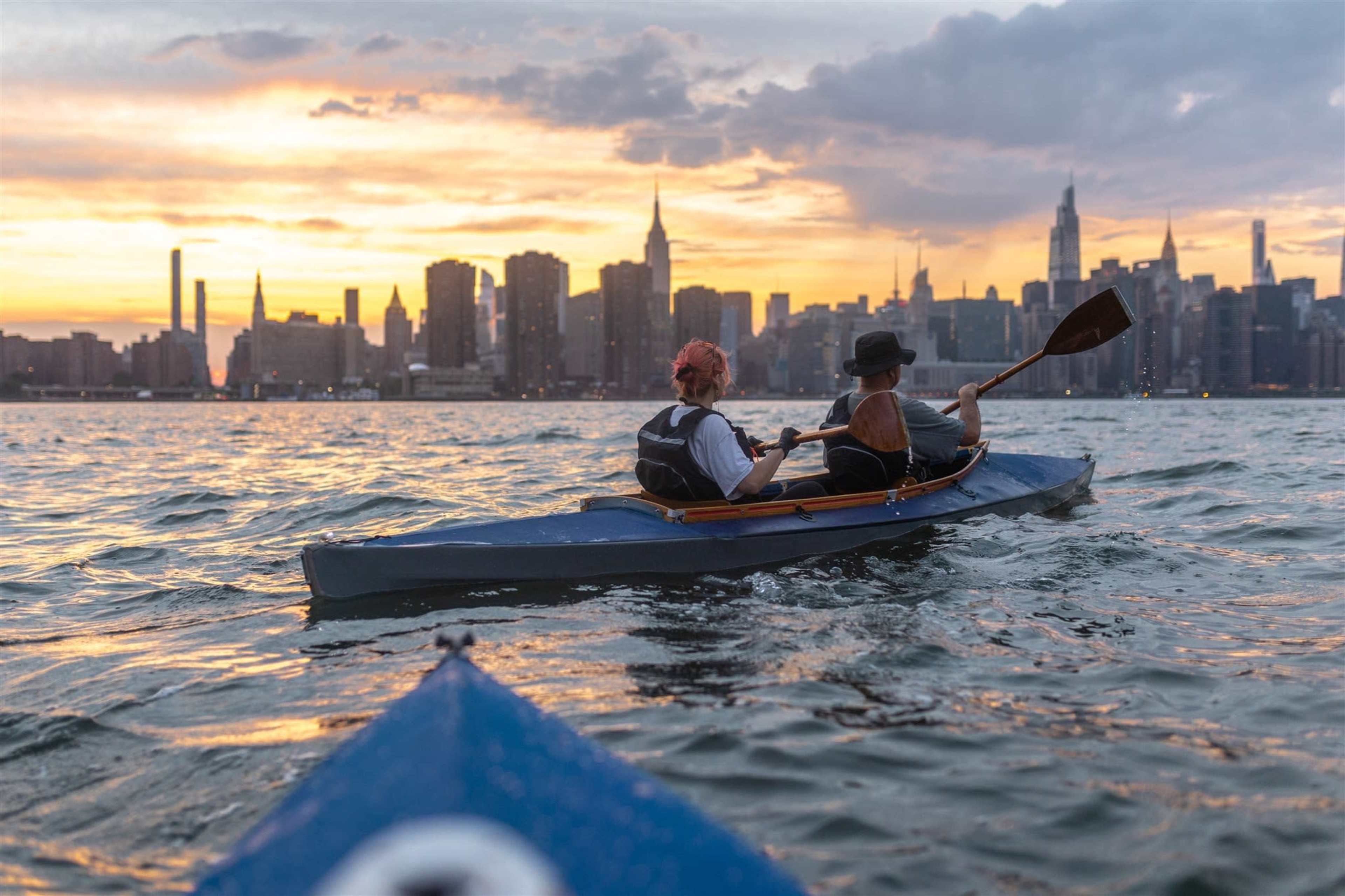 Two people are kayaking on a river with a city skyline and a sunset in the background.