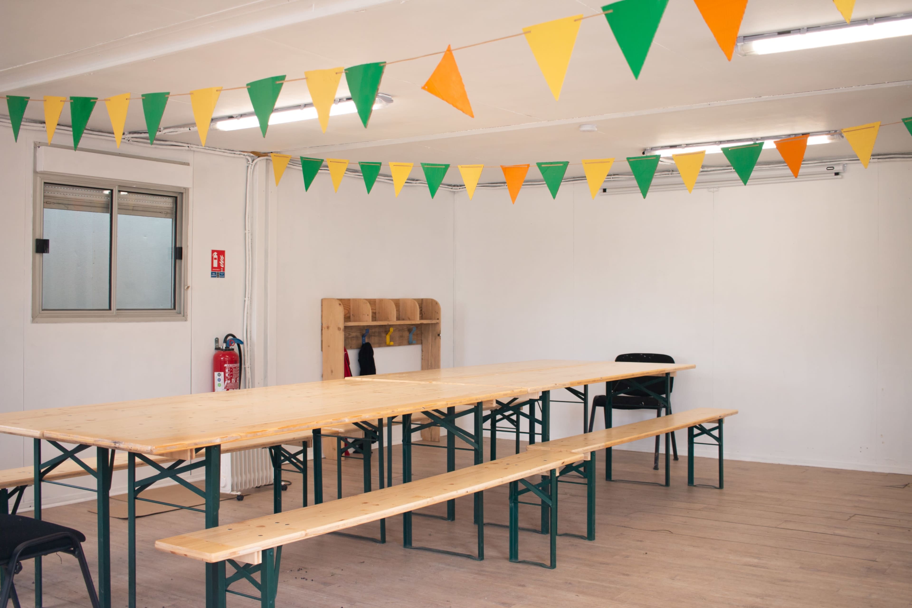 A spacious room features a long wooden table with benches and decorative green and orange bunting overhead.