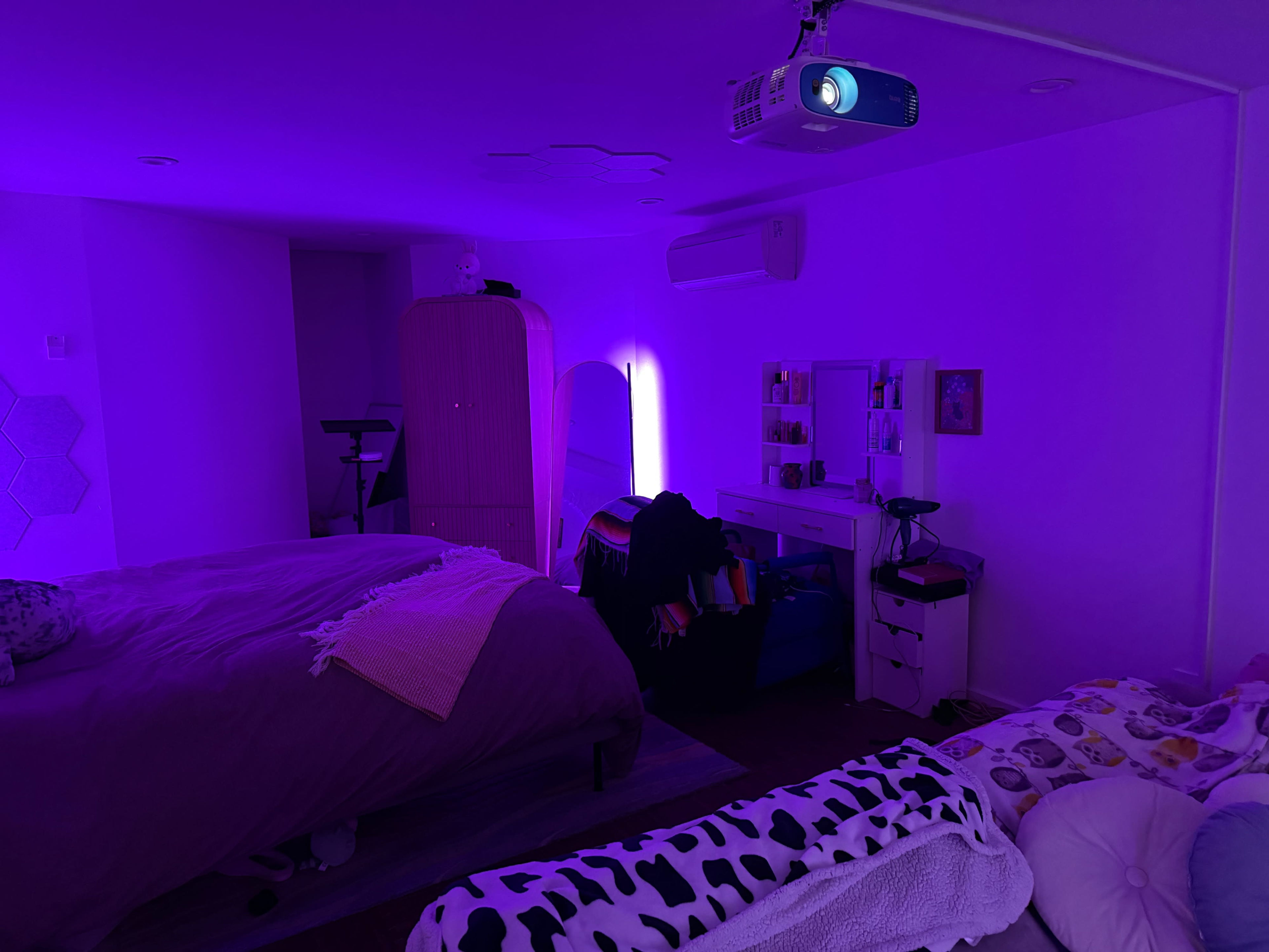 The image shows a dimly lit bedroom with purple lighting, two beds, a dresser, and a projector mounted on the ceiling.