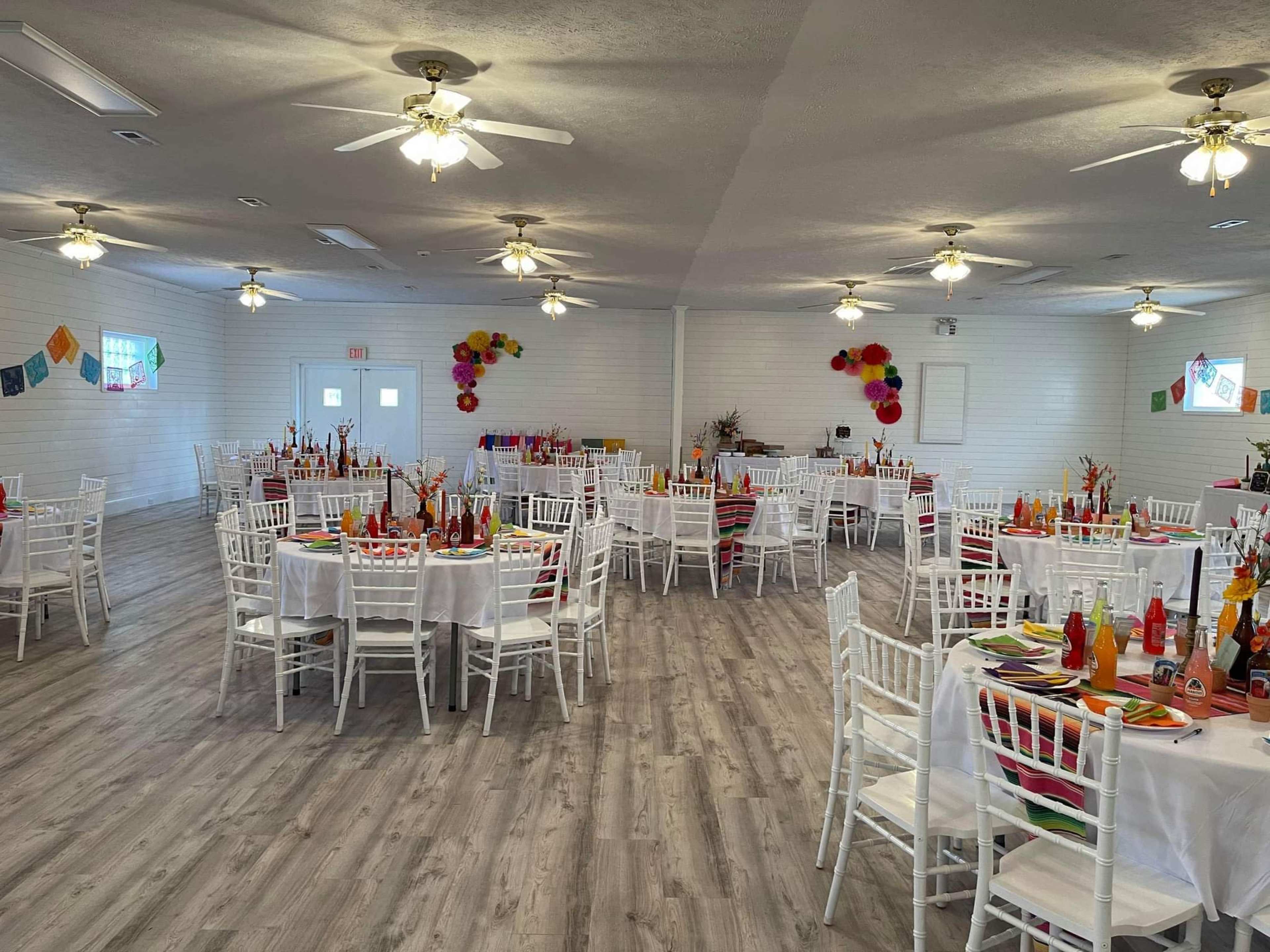 A spacious banquet hall is arranged with multiple round tables covered in white tablecloths and surrounded by white chairs, decorated with colorful centerpieces and banners.