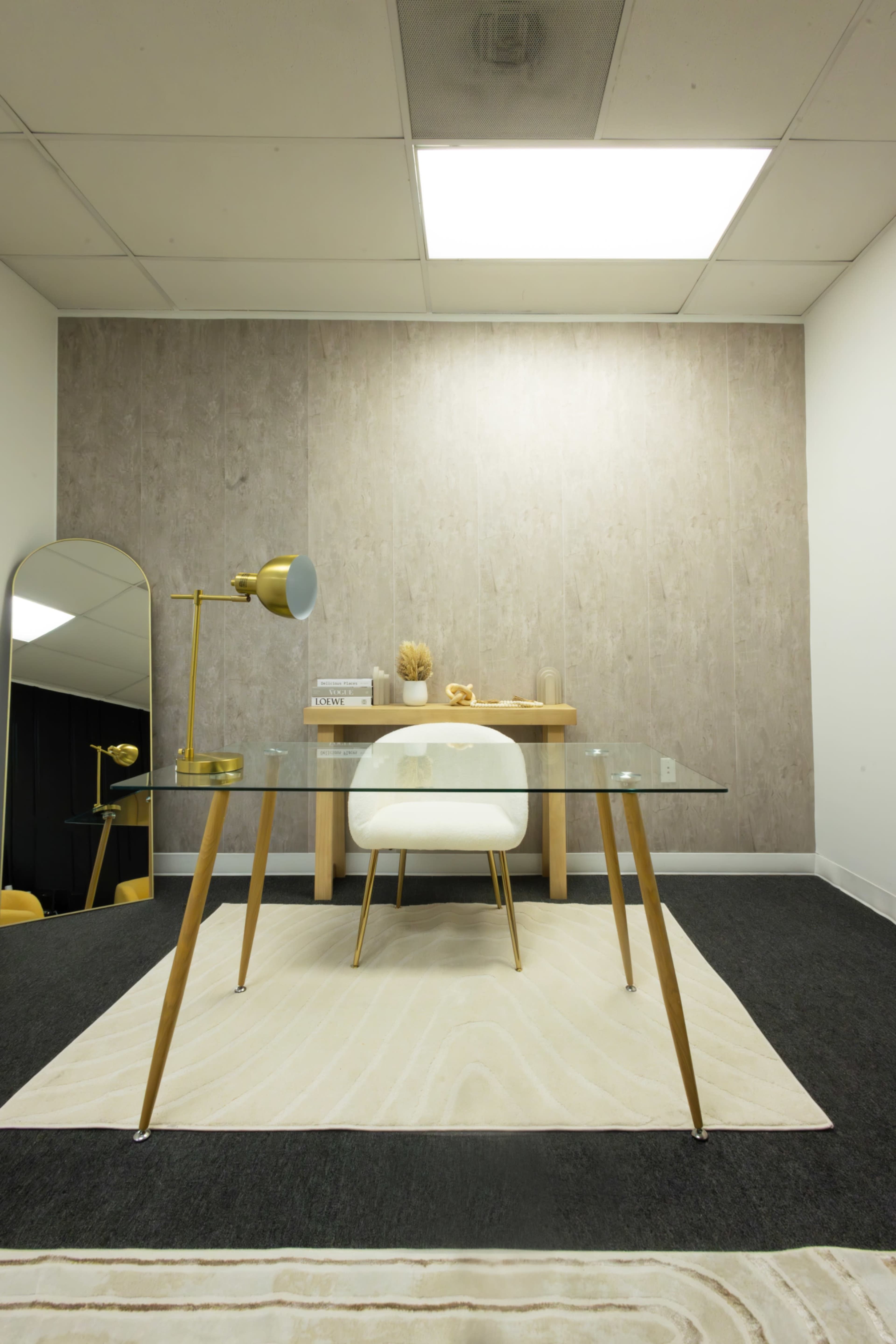 The image shows a modern office space with a glass desk, a white chair, a floor lamp, and a decorative mirror against a neutral-colored wall and carpet.