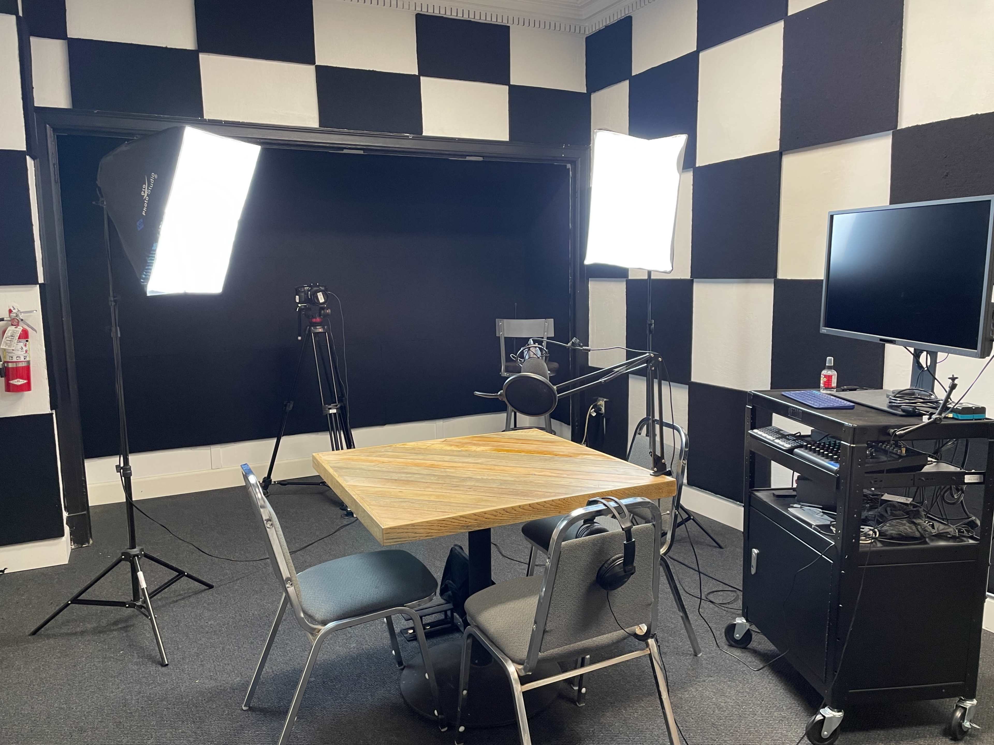 A room set up for video recording, featuring a wooden table, two chairs, studio lights, a microphone on a stand, and a computer workstation against a checkered wall.