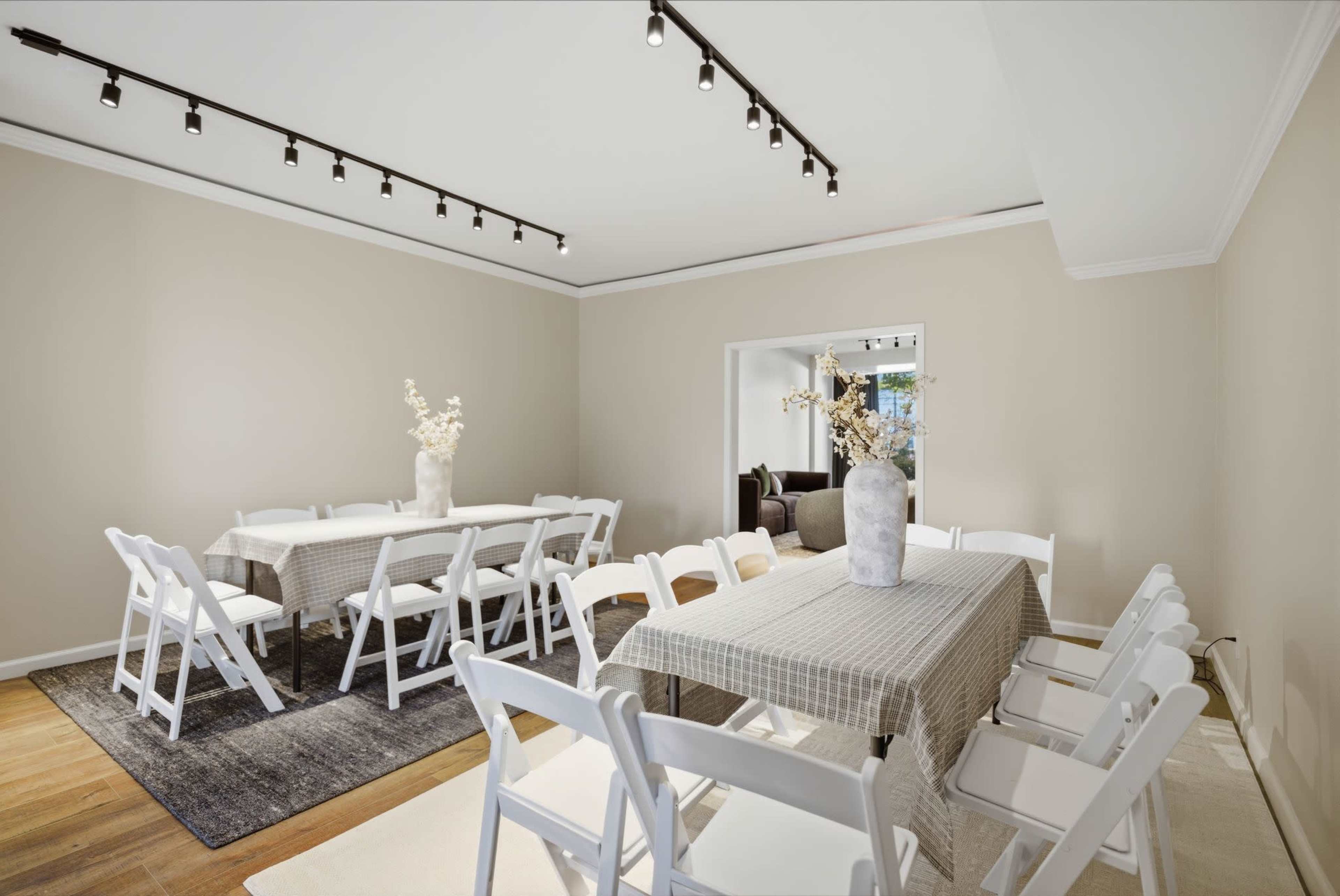 A spacious dining area features multiple white folding tables and chairs, with a decorative vase on a central table and soft lighting from track fixtures above.