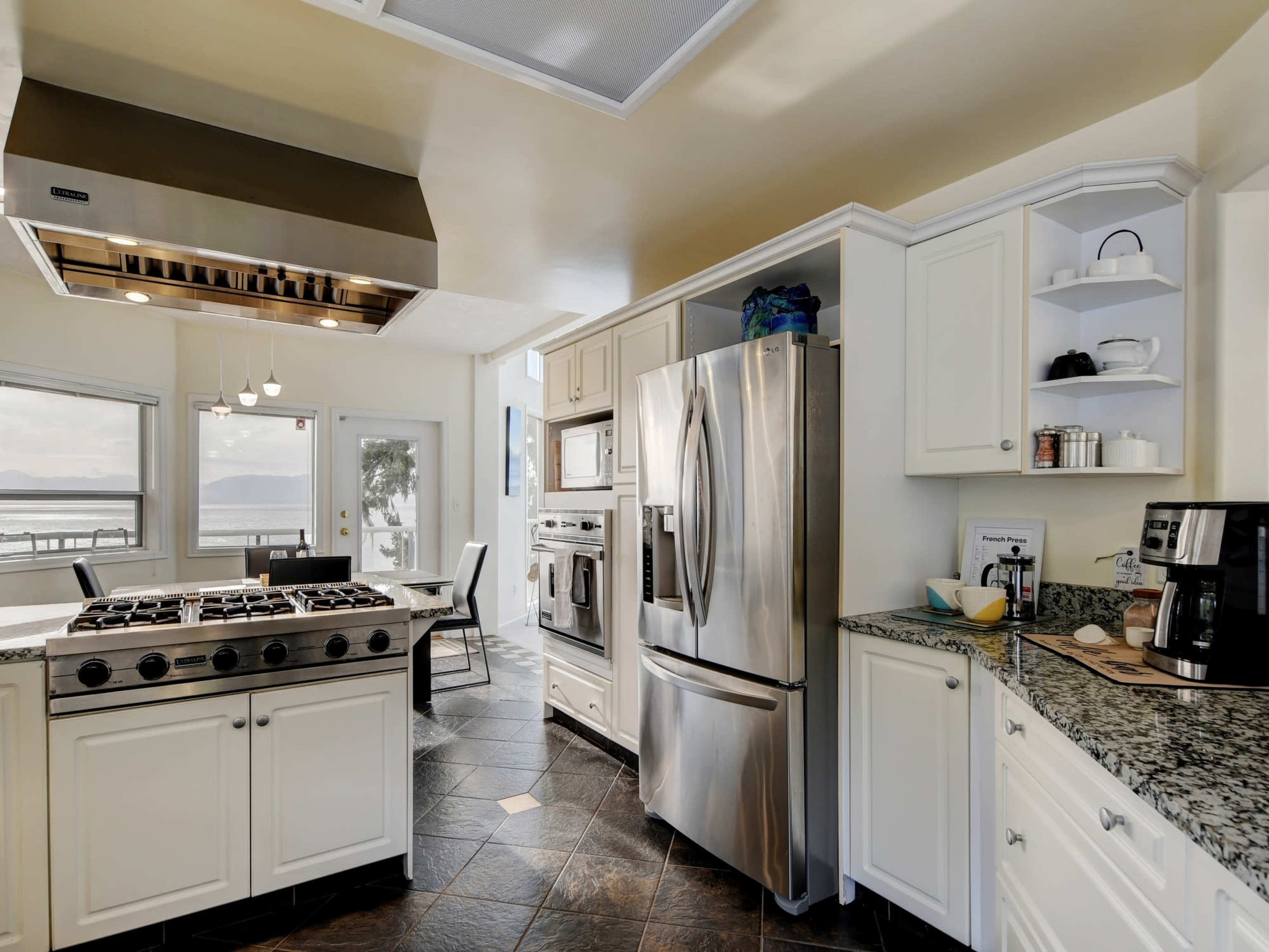 A modern kitchen features stainless steel appliances, white cabinetry, and a granite countertop with a view of the water through large windows.