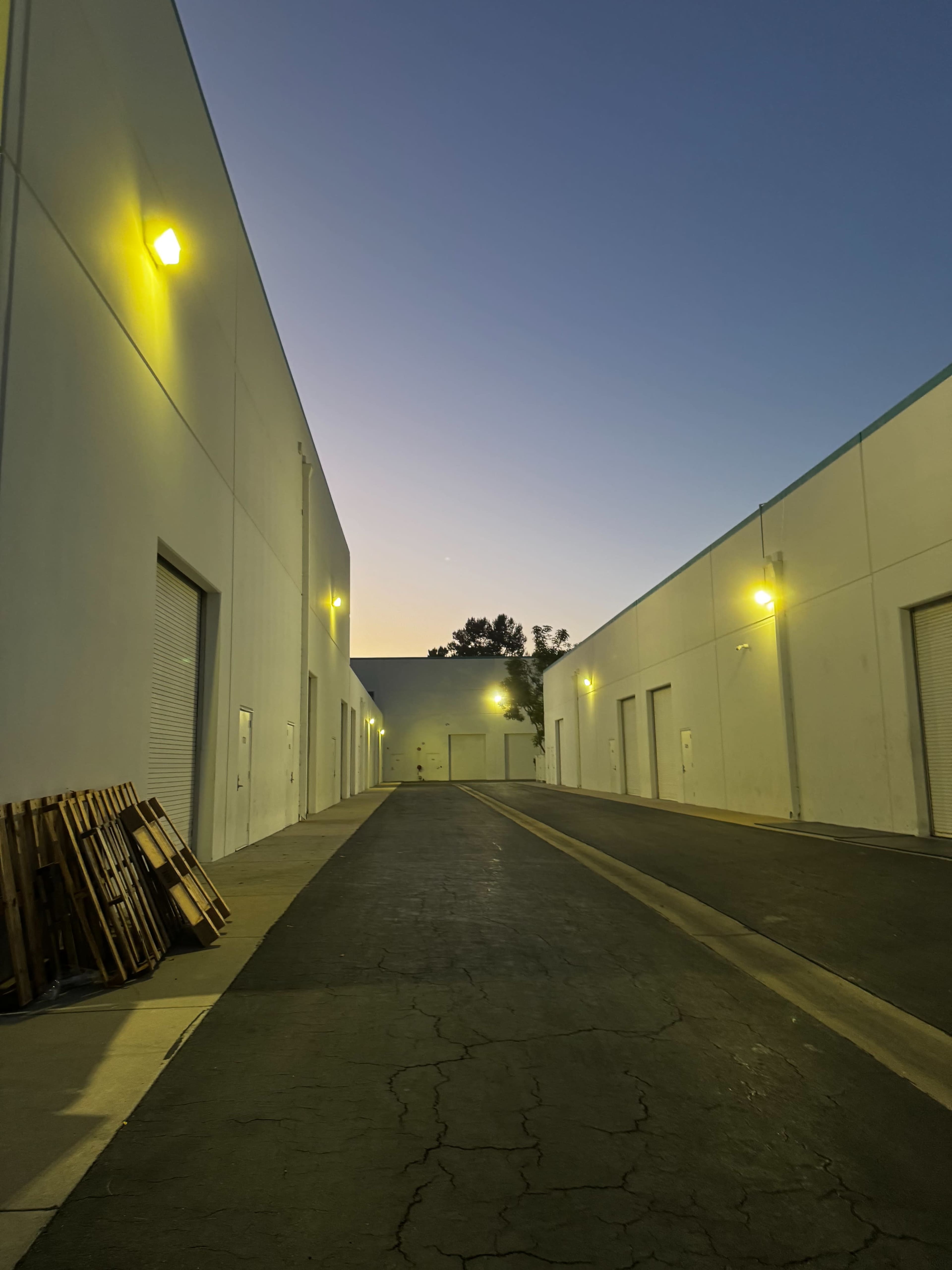 The image shows a narrow alleyway between two industrial buildings at dusk, with lighting illuminating the path and wooden pallets stacked to one side.