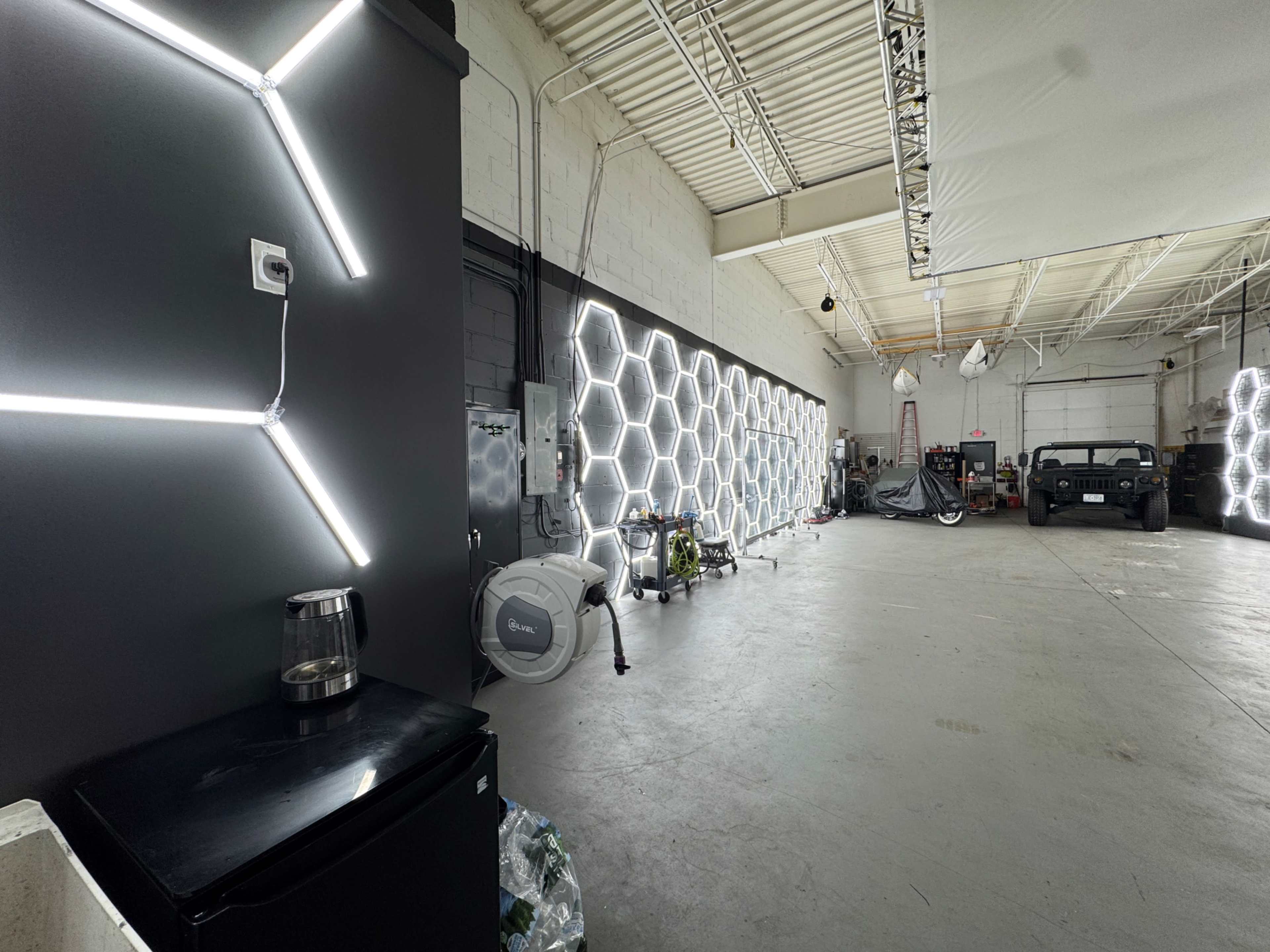 The image shows a spacious, industrial-style interior featuring a black wall with modern lighting and a row of hexagonal light fixtures along one side, with various equipment and a parked vehicle in the background.