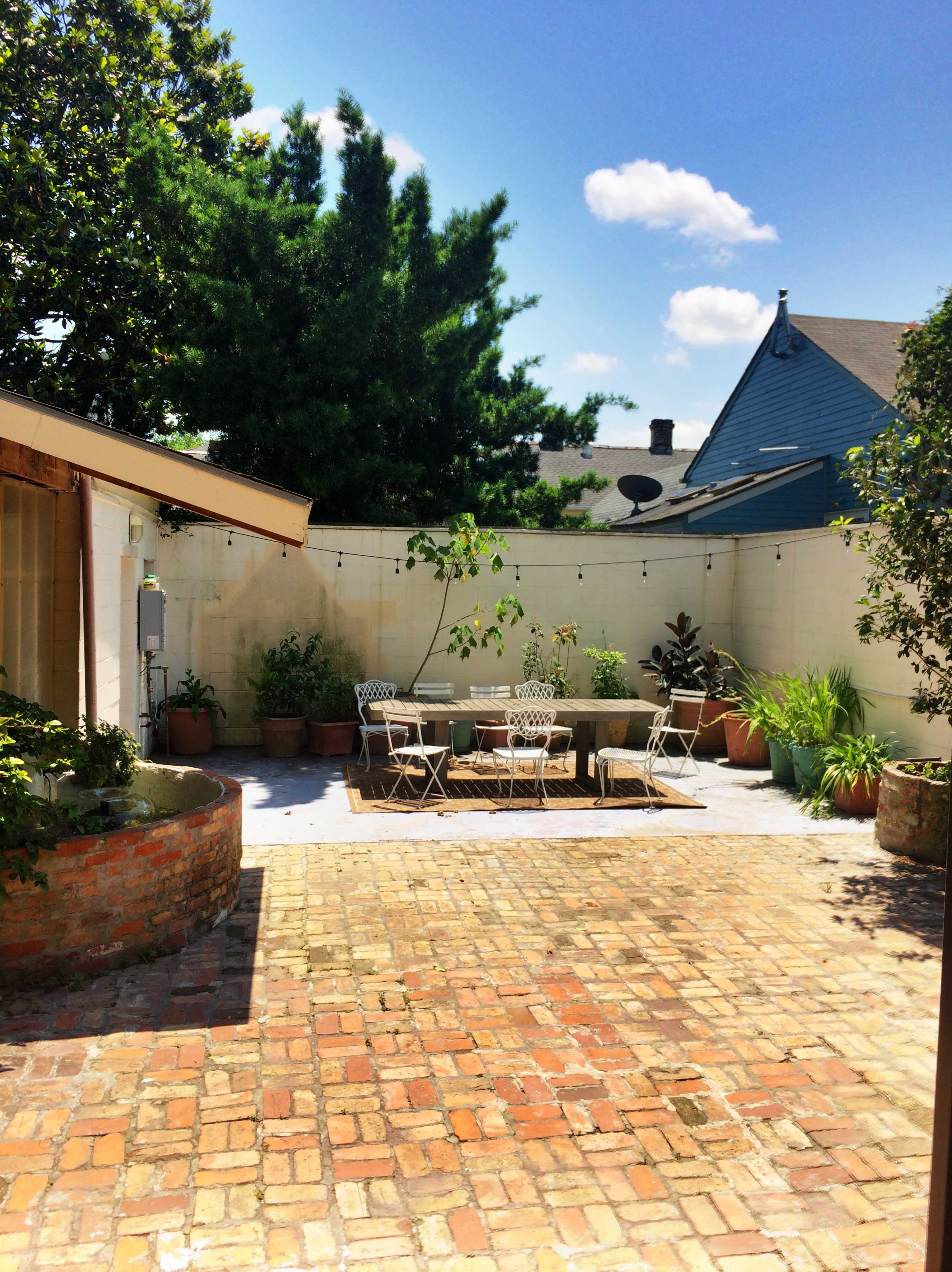 A brick-paved patio features a circular table and chairs surrounded by potted plants and a wall with a green vine.