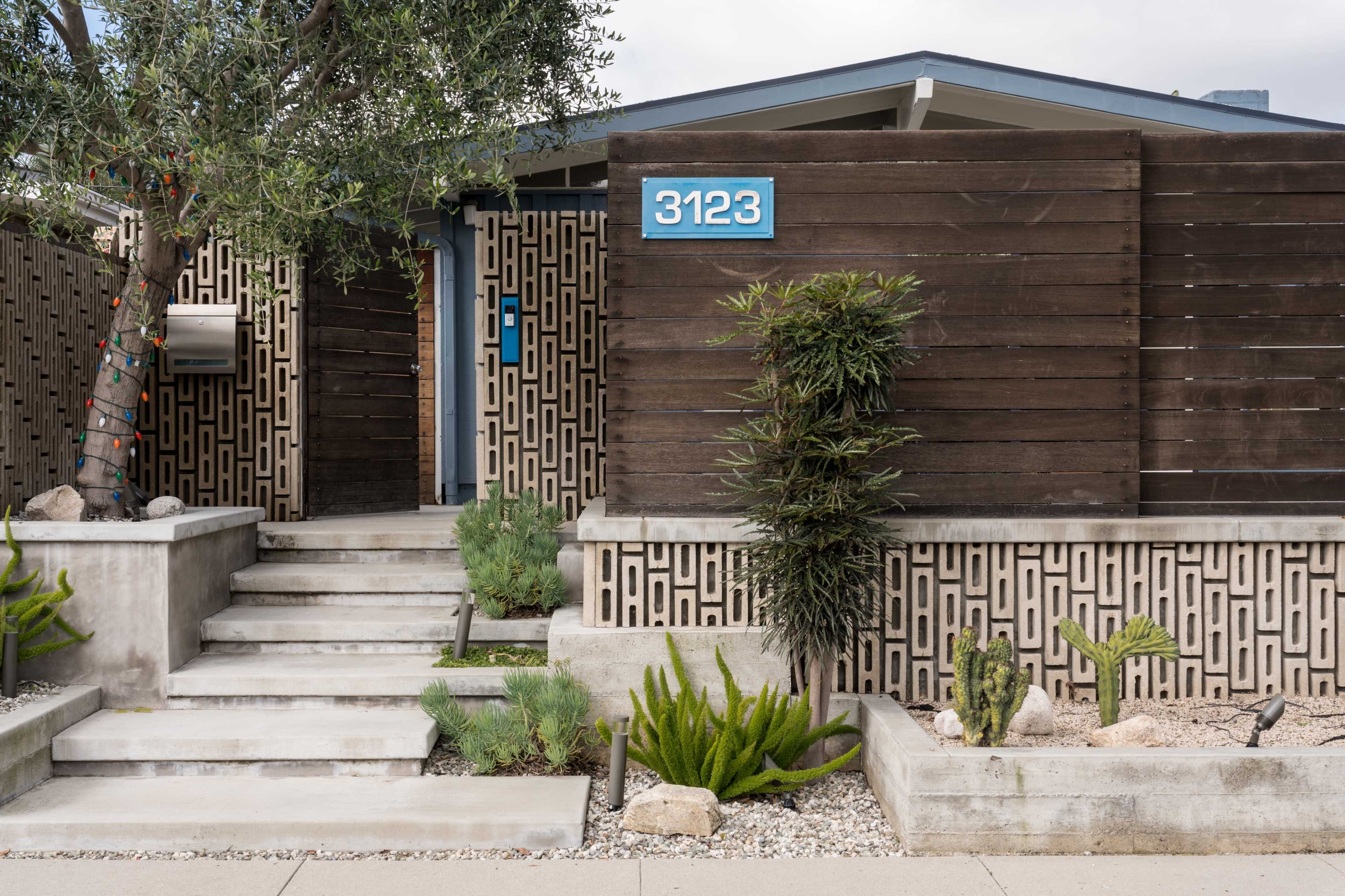 The image shows a modern home entrance featuring a wooden fence, stone steps, and various green plants in a landscaped area.