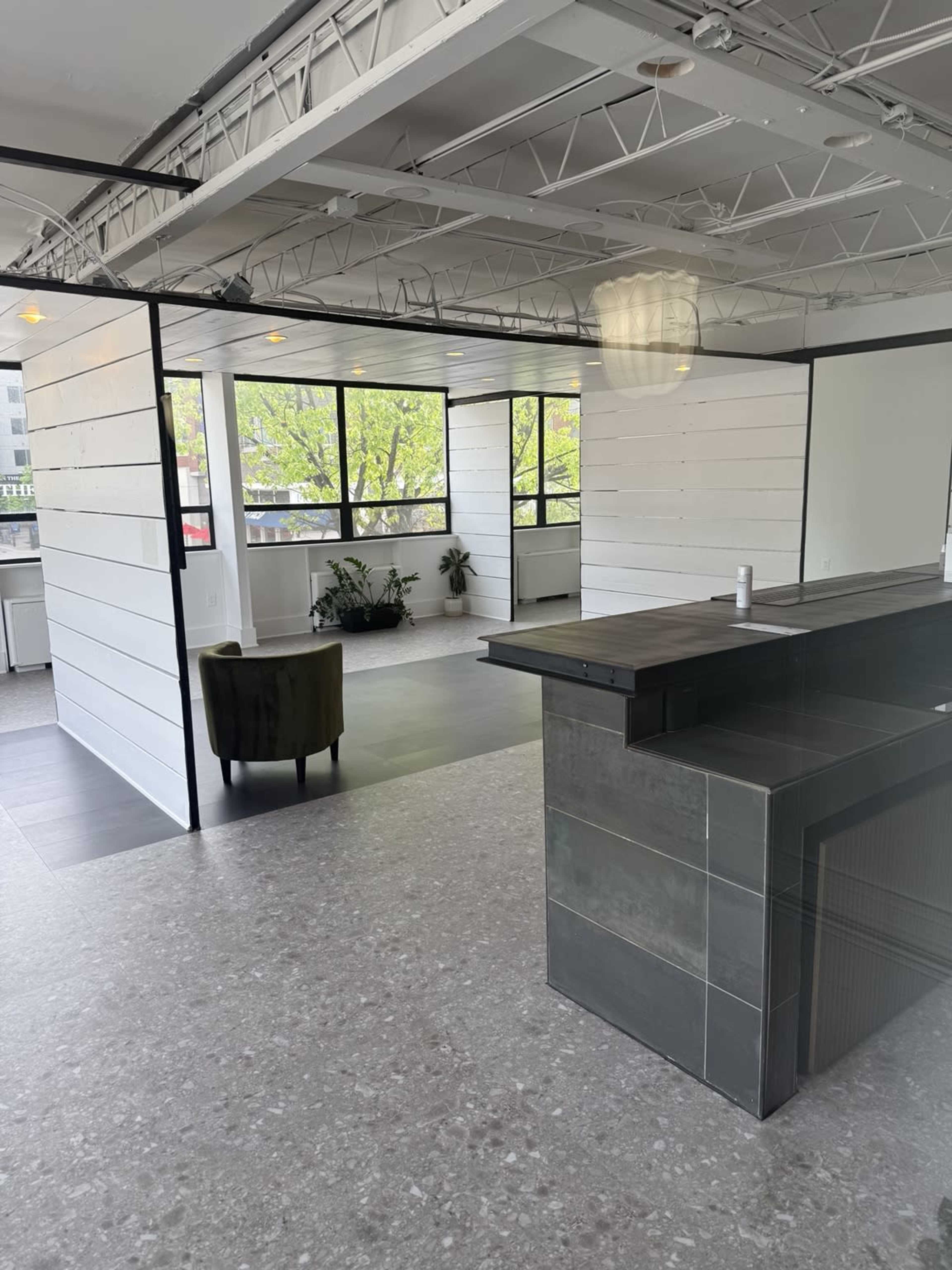 The image shows an open office space with large windows, a dark countertop, a green armchair, and a minimalist aesthetic featuring white walls and industrial elements.