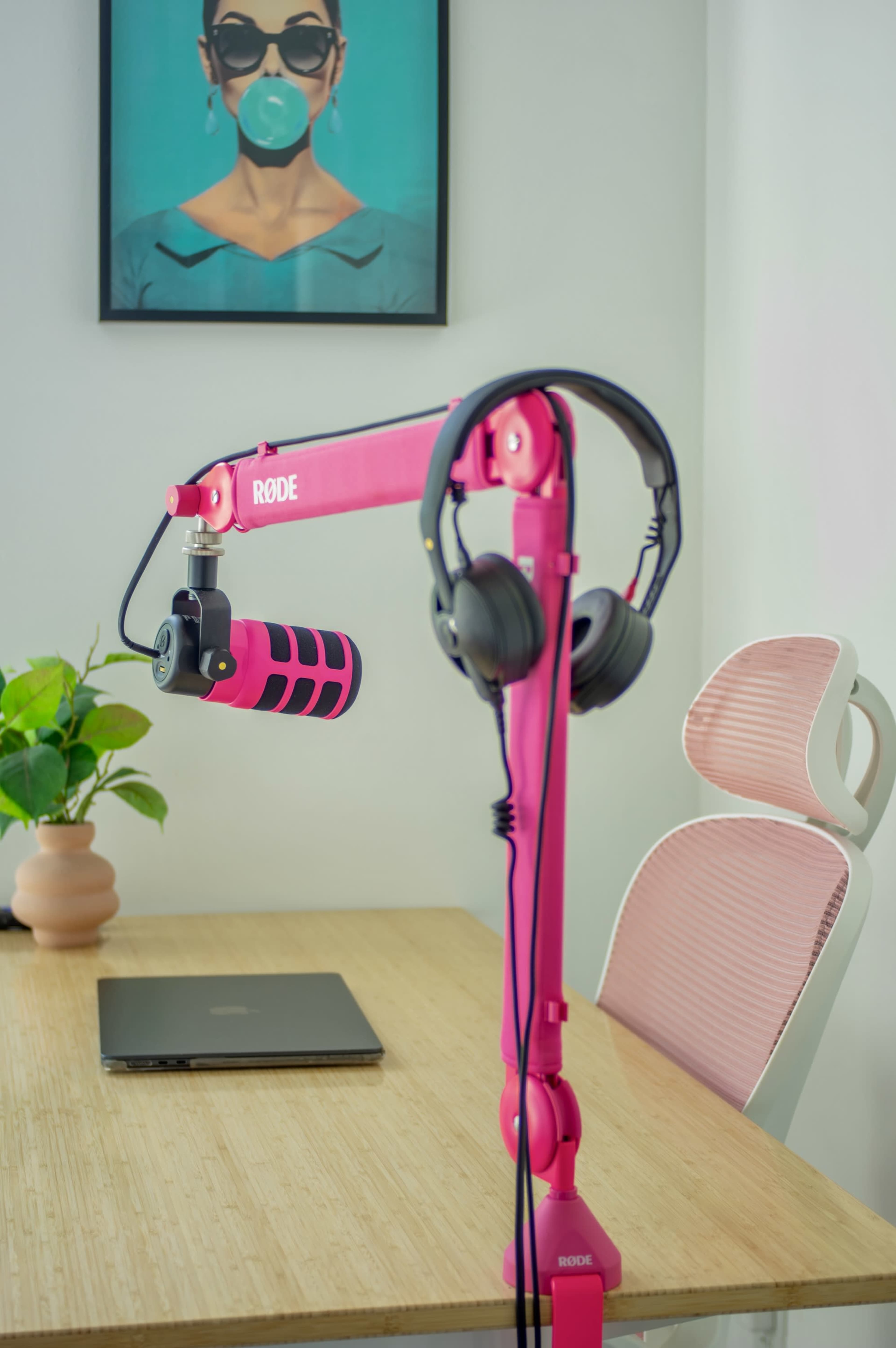 A pink microphone on a stand with headphones attached is positioned next to a laptop on a wooden table, beneath a framed portrait on the wall.