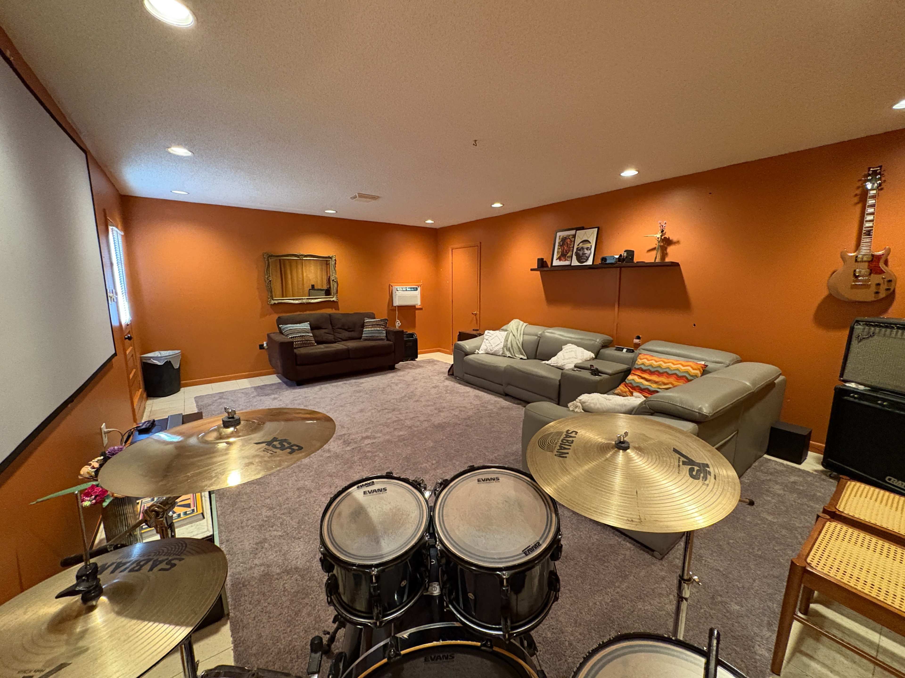 The image shows a spacious basement room with orange walls, a drum set in the foreground, and various seating arrangements including couches and a chair.
