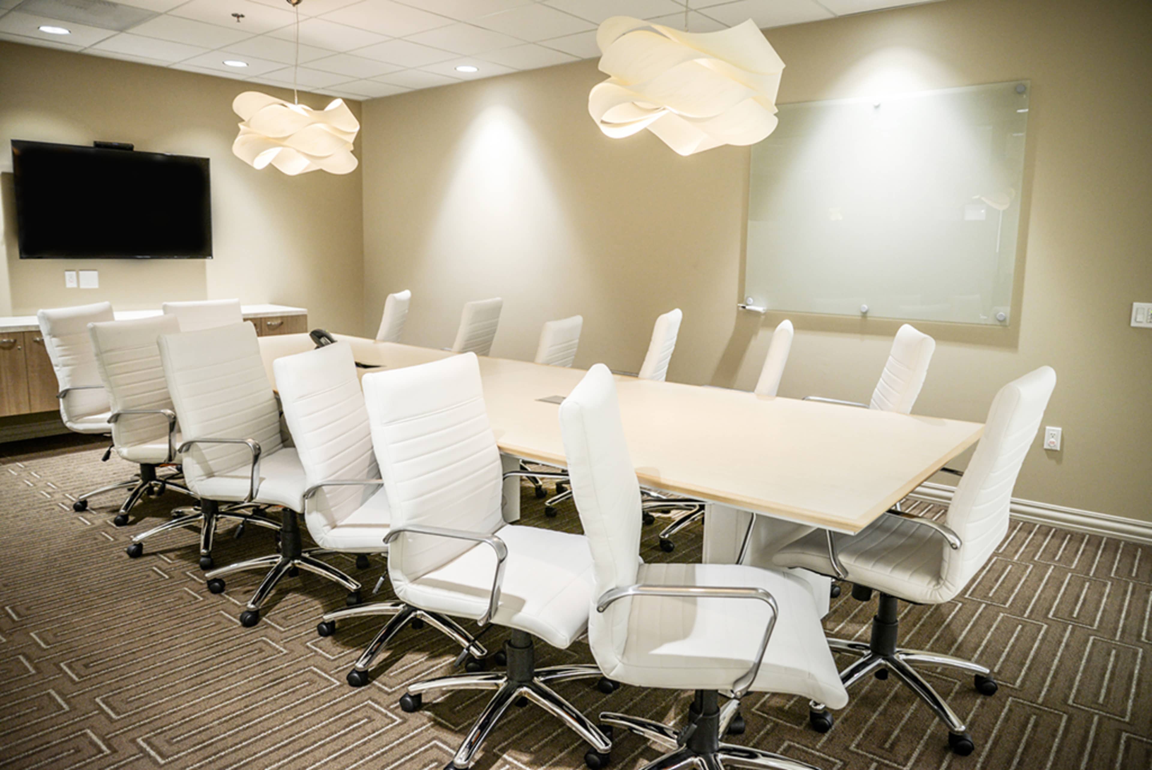 A modern conference room is set up with a long table surrounded by white ergonomic chairs under decorative light fixtures.