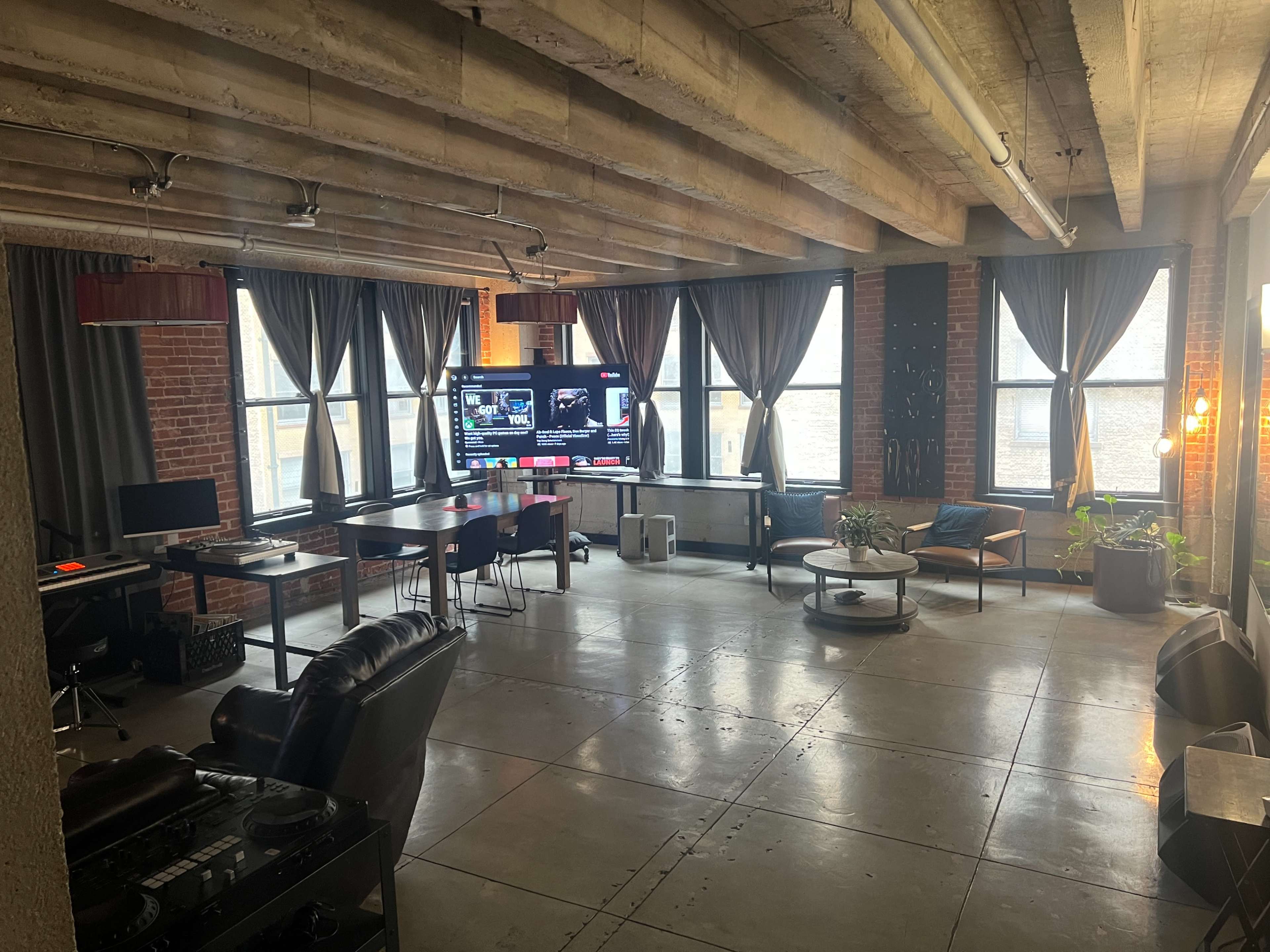 The image shows a spacious room with exposed brick walls, large windows, a mix of work and lounge furniture, and a large screen displaying content on one side.
