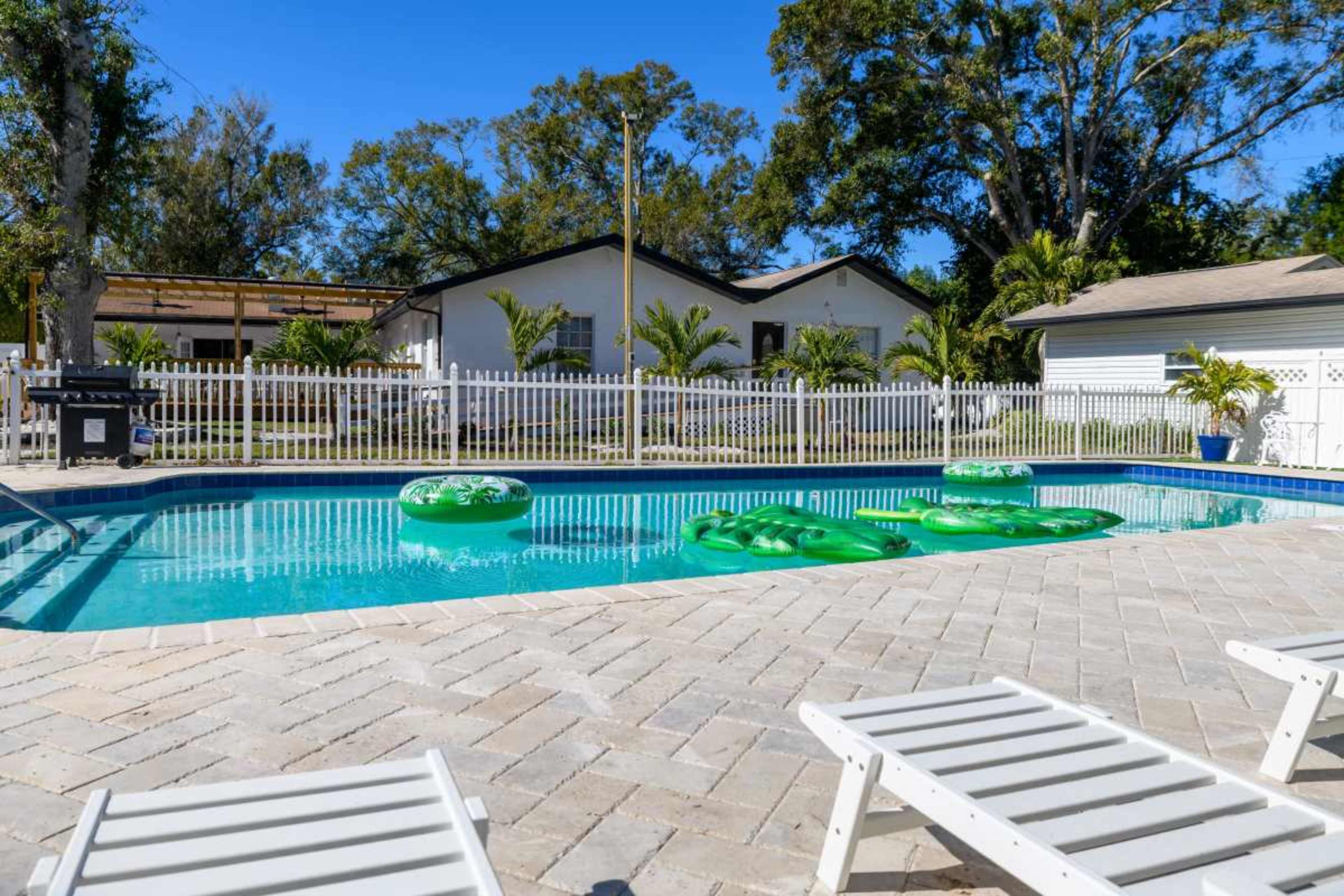 The image shows a swimming pool surrounded by lounge chairs, with green floatation devices in the water and a white fence in the background.