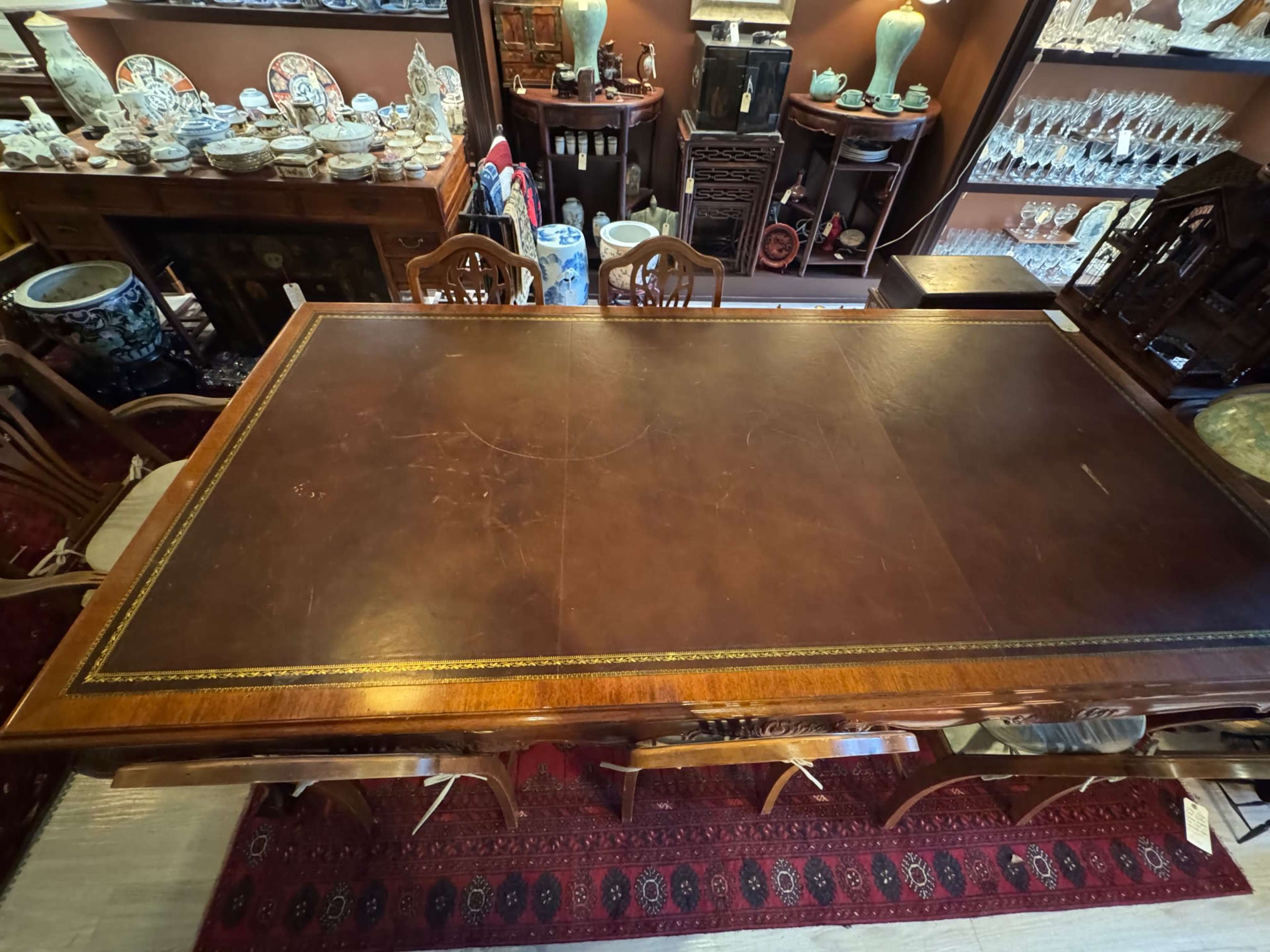 A large wooden table with a leather top is displayed in a room filled with various antique items and decorative glassware.