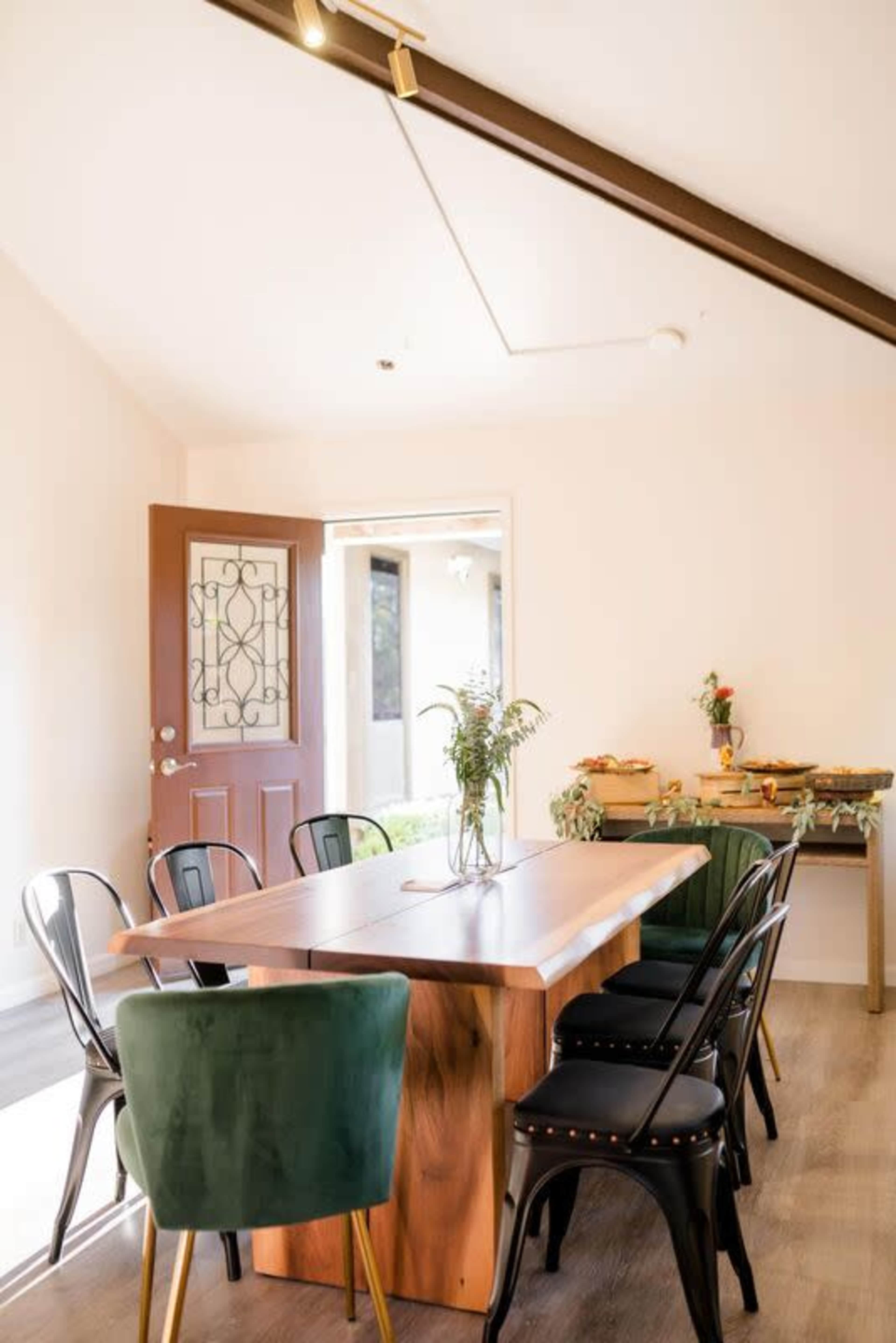 A wooden dining table with green and black chairs is positioned in a well-lit room with an open door leading to an outdoor area.