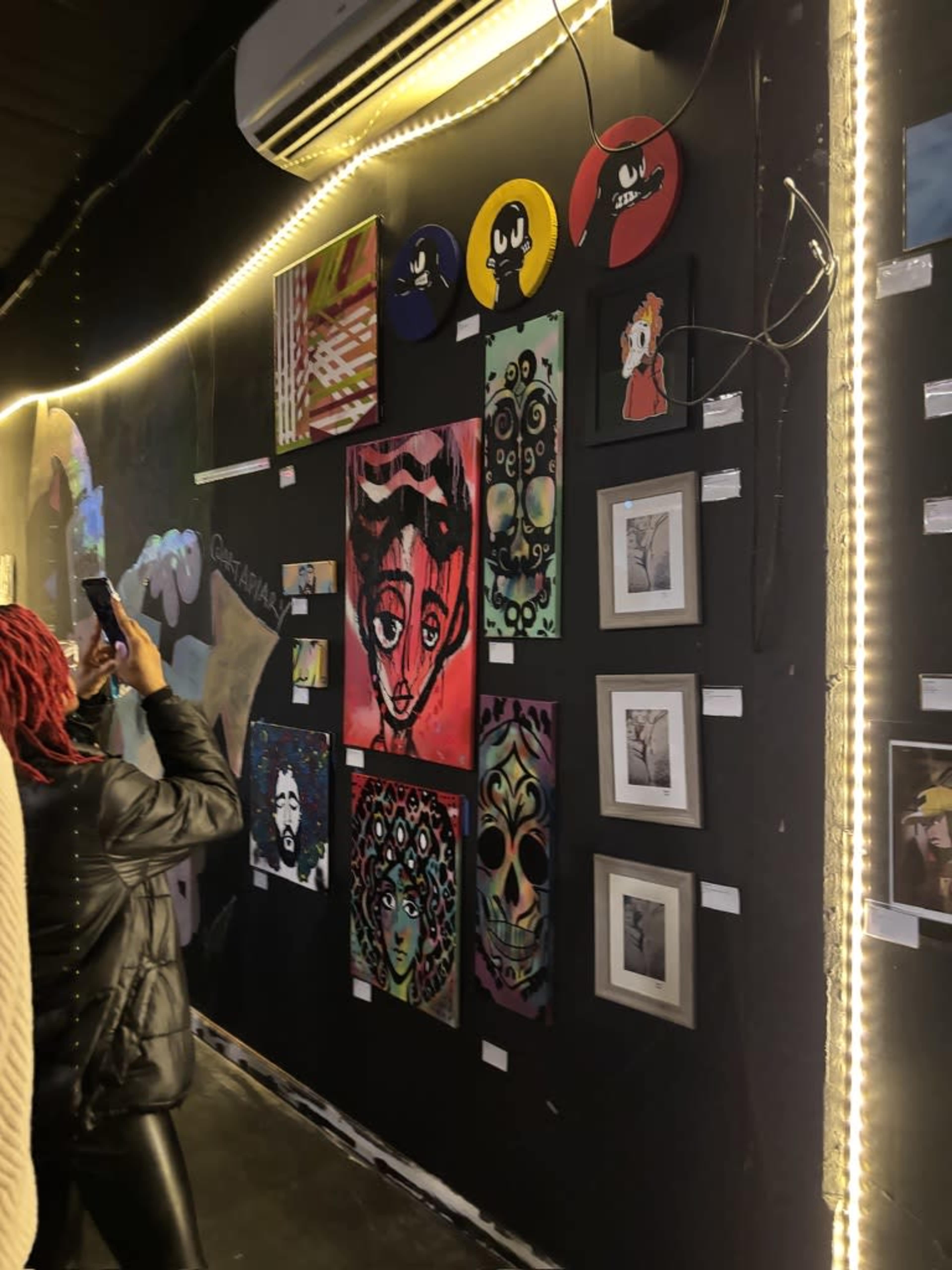 The image shows a gallery wall displaying a variety of colorful artworks alongside a visitor taking a photograph.