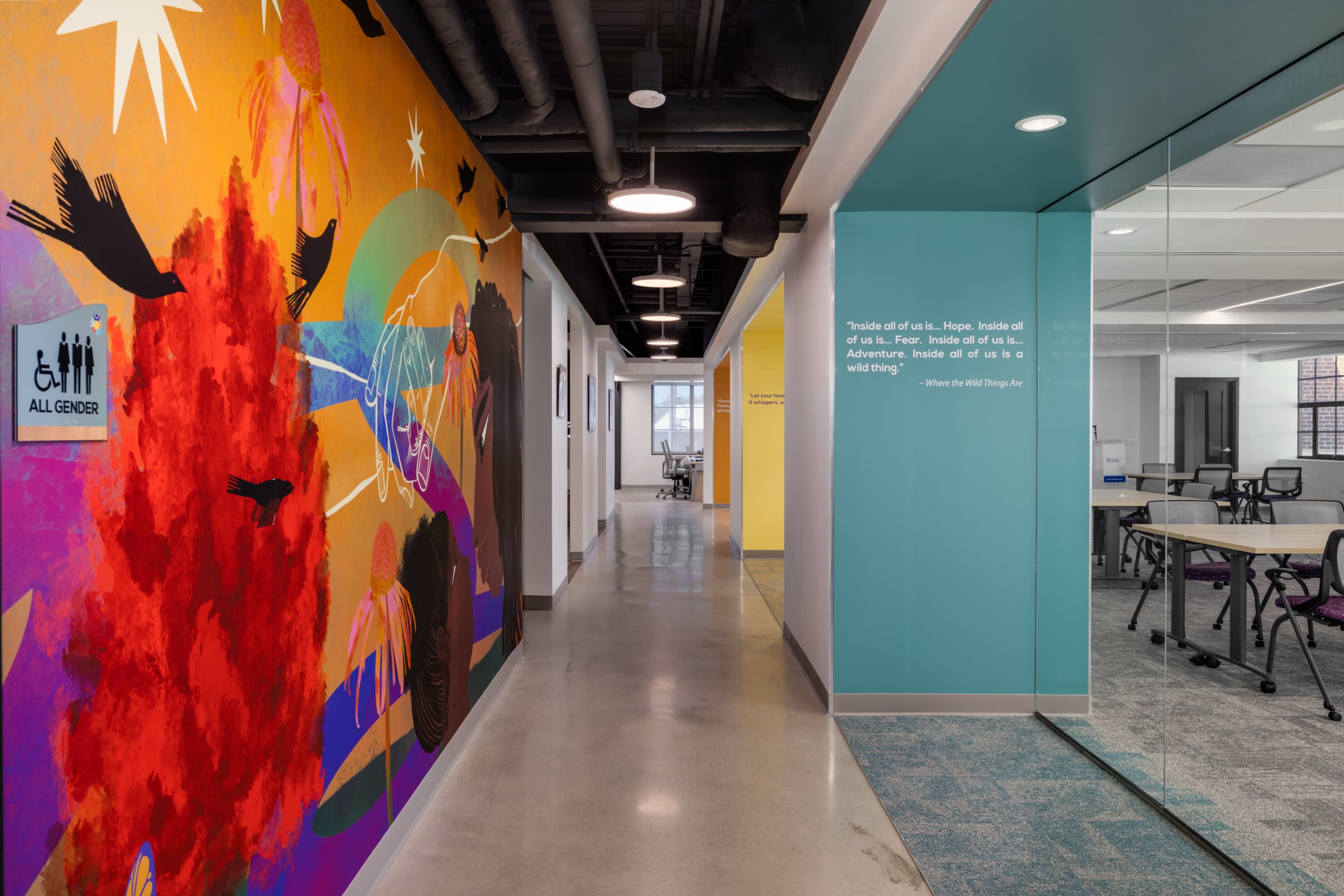 The image shows a vibrant hallway featuring a colorful mural on one wall, with a sign indicating all-gender restrooms and glass office spaces visible in the background.