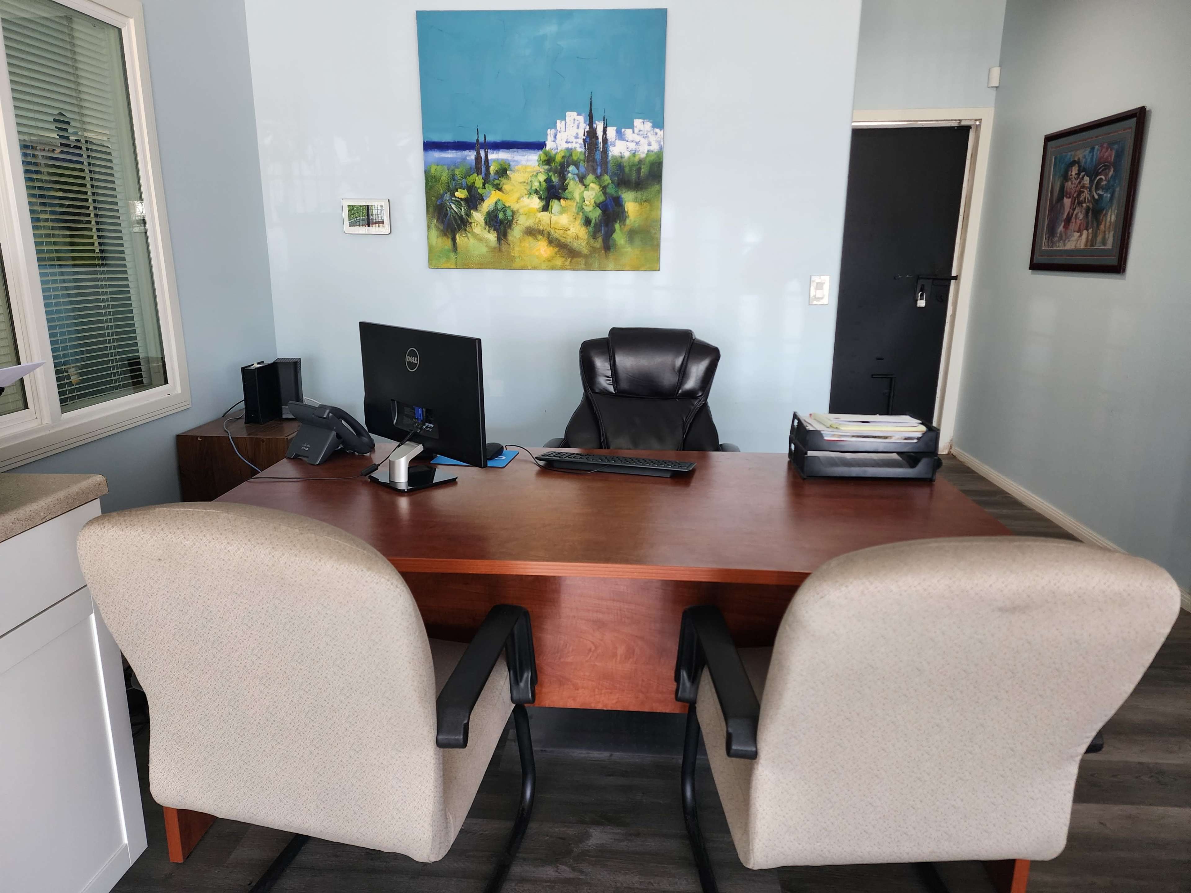 The image shows a modern office with a wooden desk, two ergonomic chairs, a computer monitor, and a large painting on the wall.
