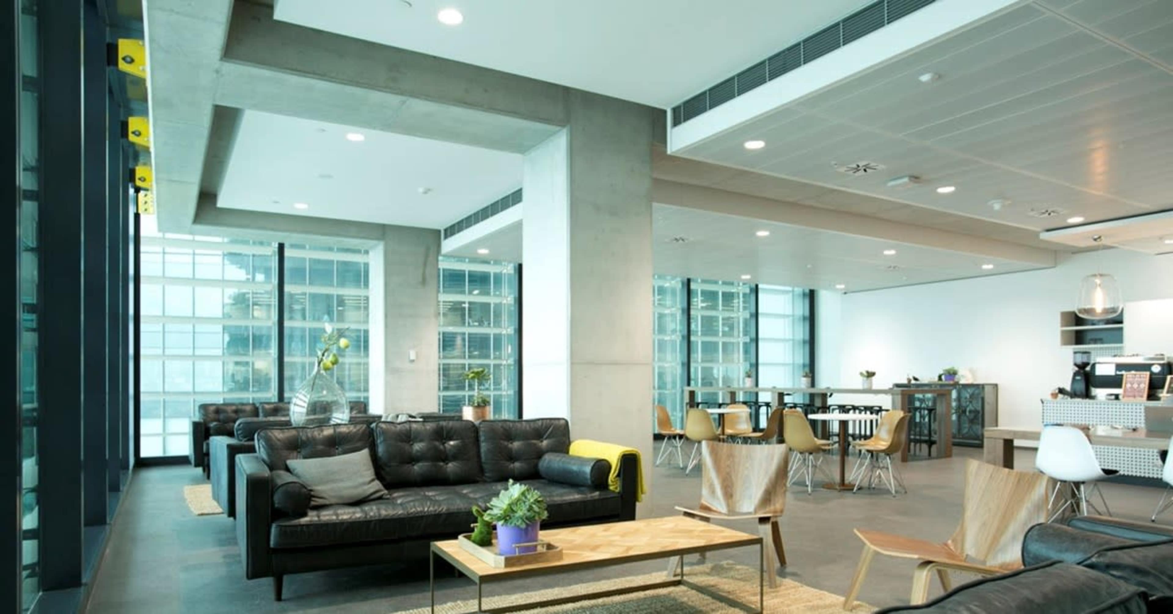 A modern office space featuring leather sofas, a wooden coffee table, and a dining area with tables and chairs, all surrounded by large windows.