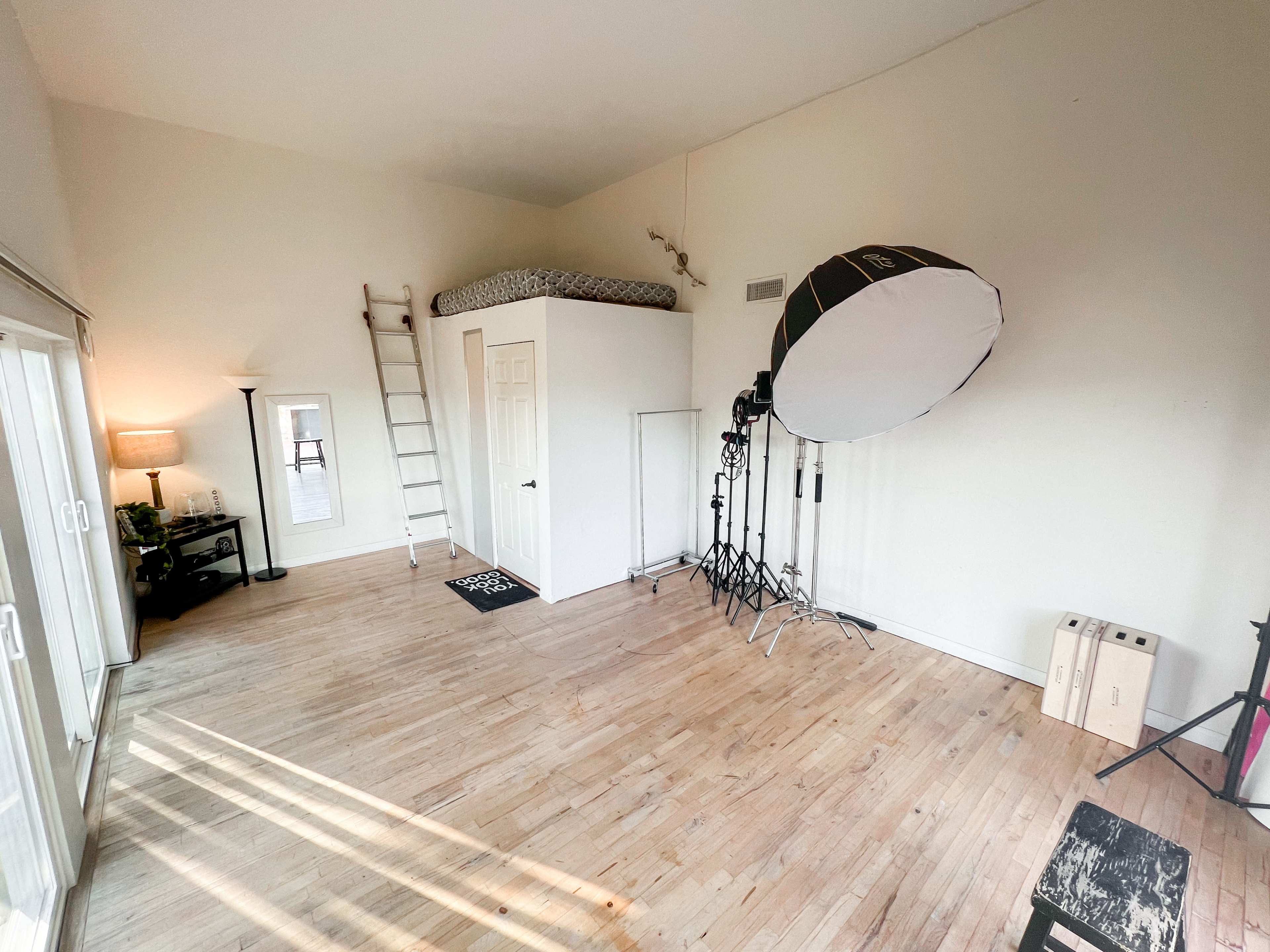The image shows a spacious room with a bed on a lofted platform, a ladder, a large light stand with a softbox, and wooden floors.